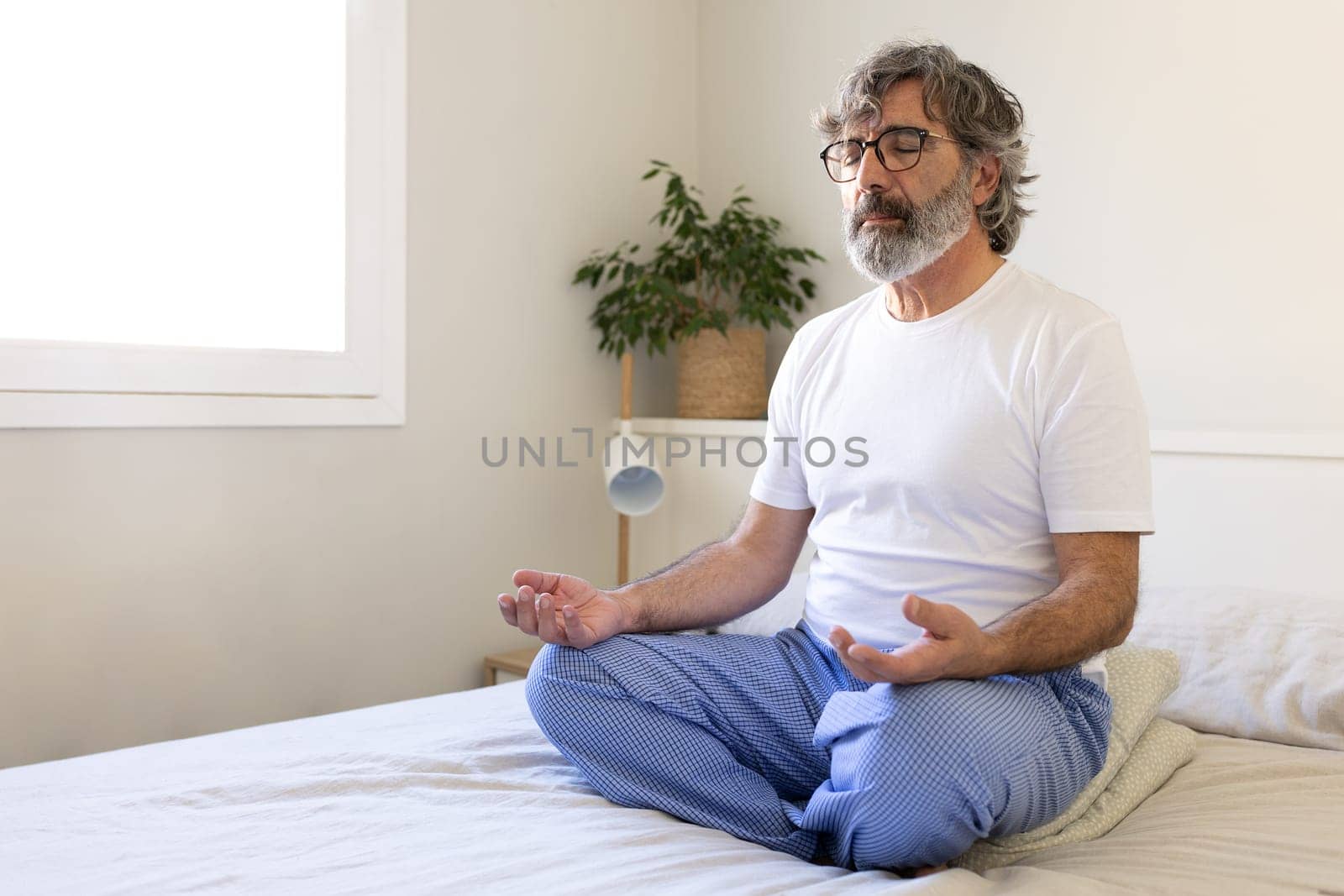 Mature adult man relaxing, doing meditation sitting on bed at home. Male meditating in the morning. Copy space. Wellness and mental health concept.