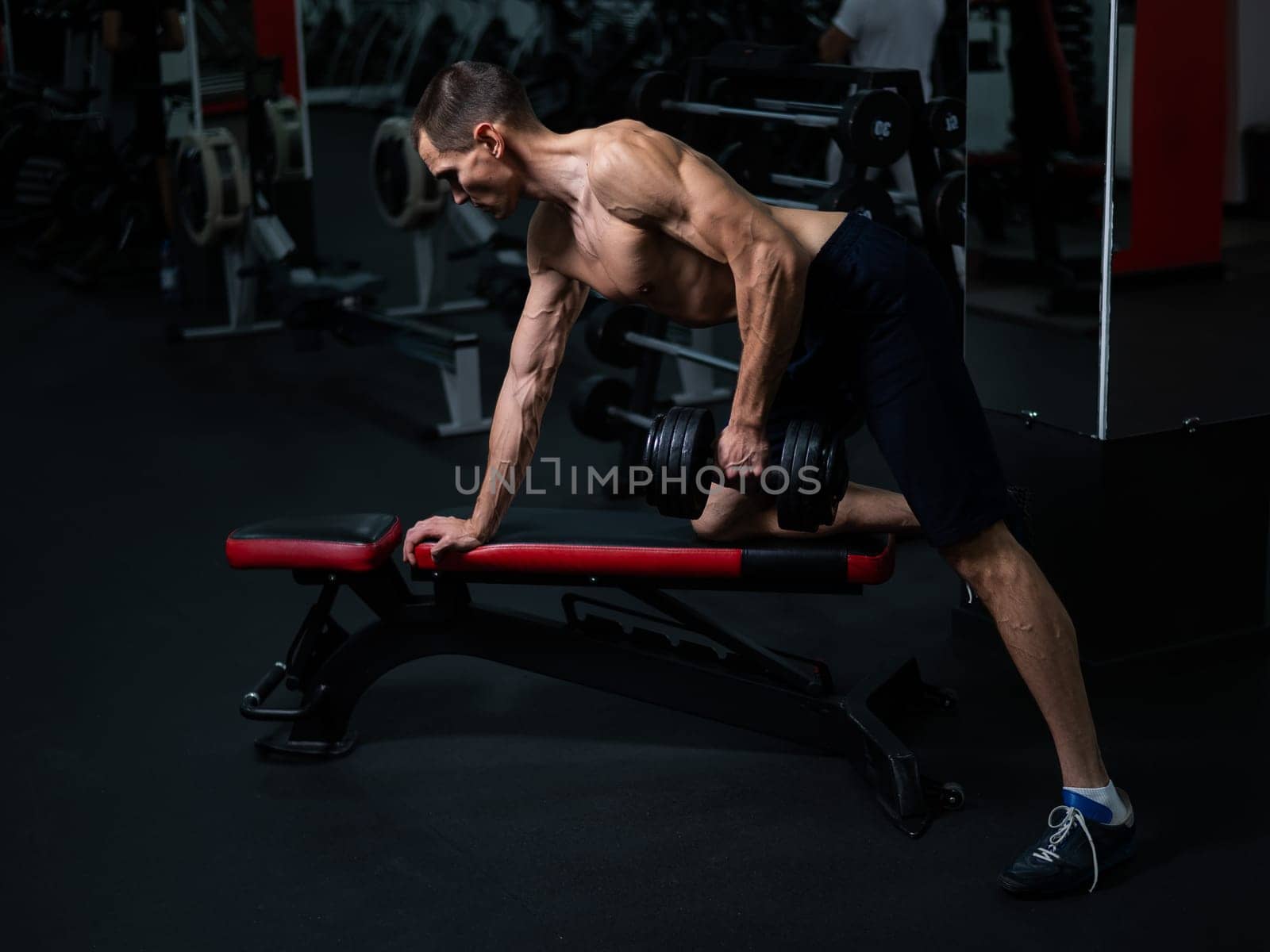 Shirtless man doing dumbbell row to waist on bench at gym