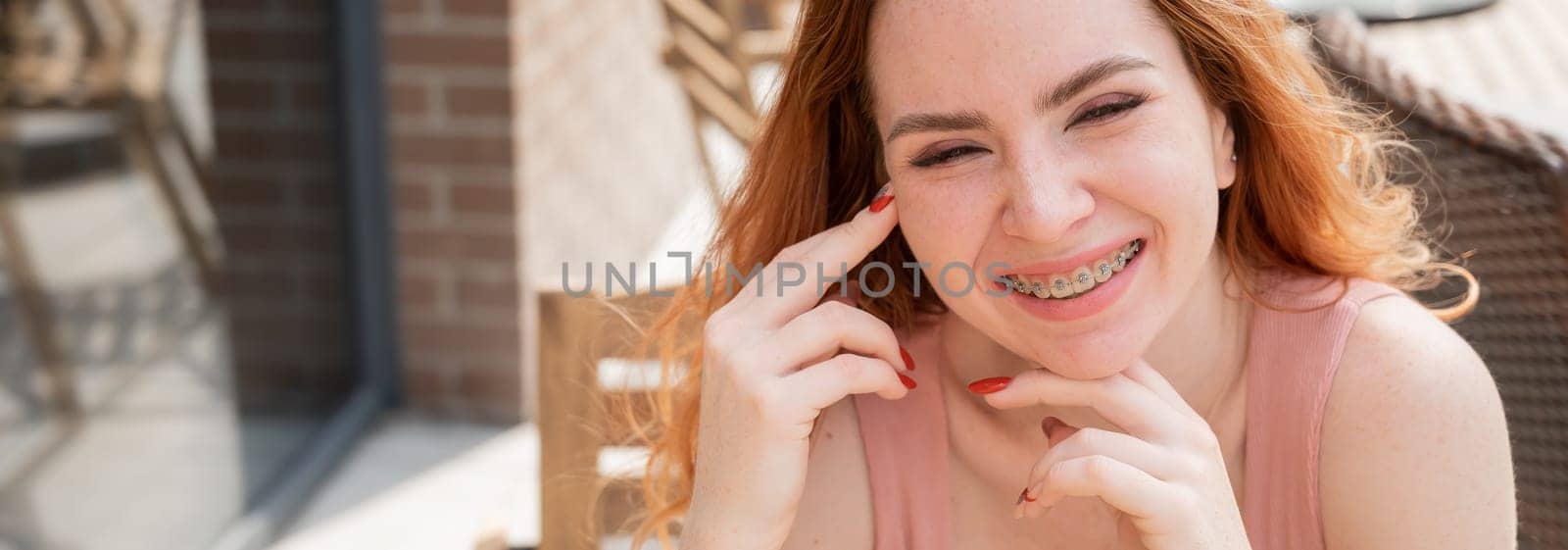 Beautiful young red-haired woman with braces on her teeth smiling while sitting in an outdoor cafe. Widescreen. by mrwed54