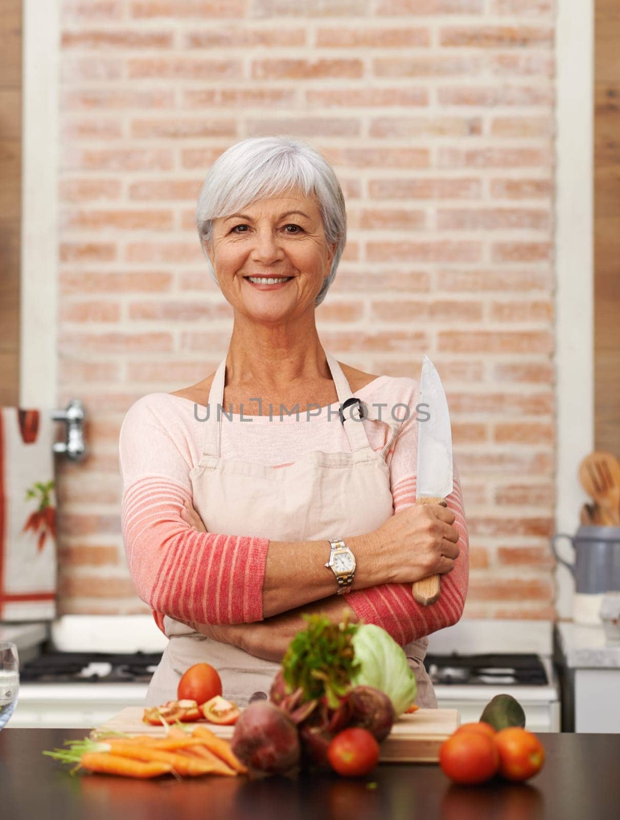 Senior woman and cooking with knife in kitchen or confident, tomato or carrots for diet. Female chef or portrait with healthy vegetables for nutrition, green lettuce and onion in retirement home by YuriArcurs