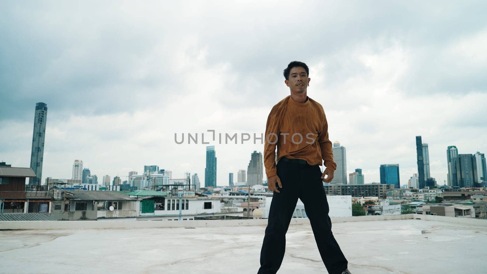 Motion shot of B-boy dance performance by professional street dancer at rooftop with sky scrapper, city view. Attractive asian hipster show energetic footstep. Outdoor sport 2024. Endeavor.