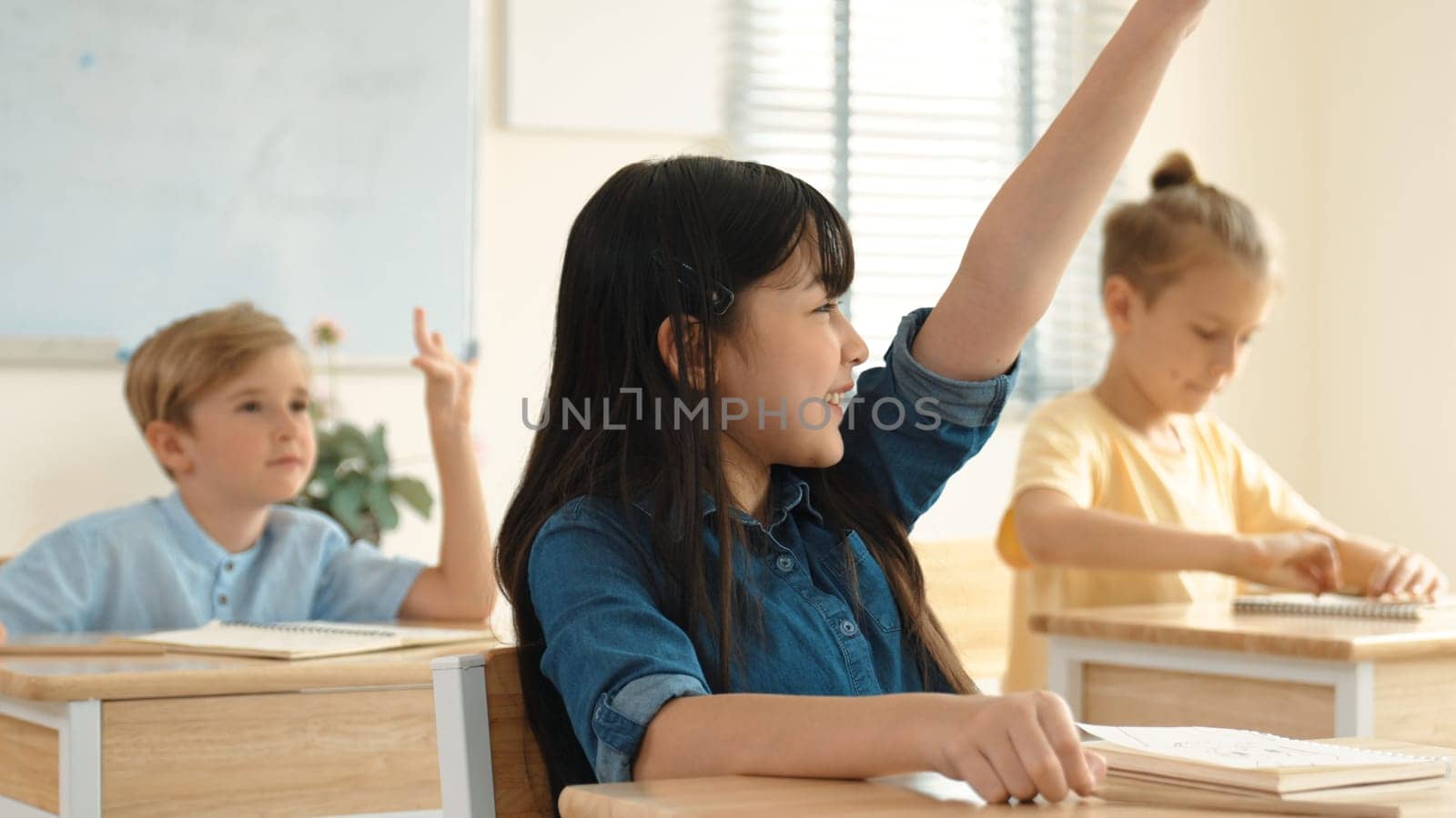 Happy girl raising hand for asking and answering teacher surrounded by multicultural children studying at classroom. Happy student working together, voting, volunteering, calling instructor. Pedagogy.