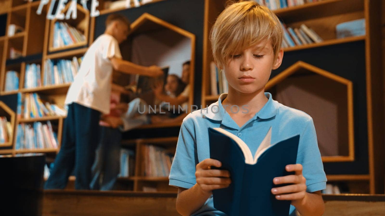 Attractive caucasian boy reading a book while group of smart students sitting at library. Child studying, learning from novel or textbook while children talking, chitchat about education. Erudition.