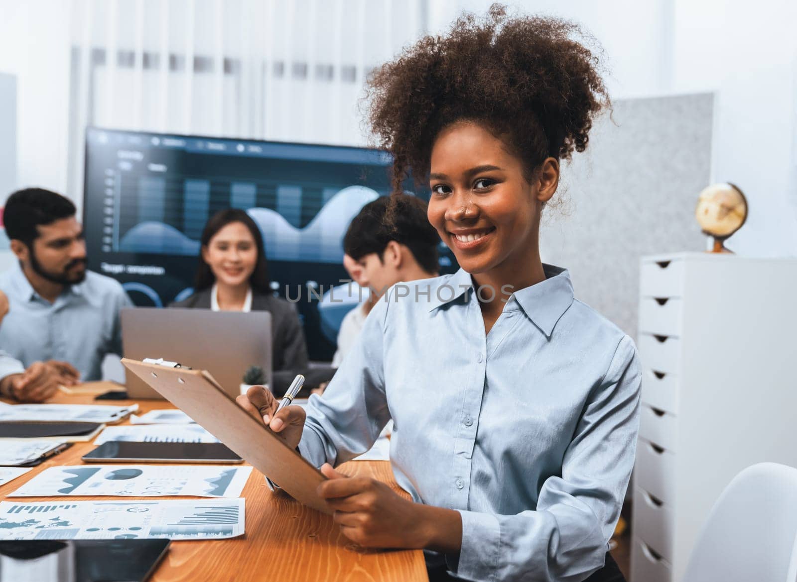 Portrait of happy young african businesswoman with group of office worker on meeting with screen display business dashboard in background. Confident office lady at team meeting. Concord