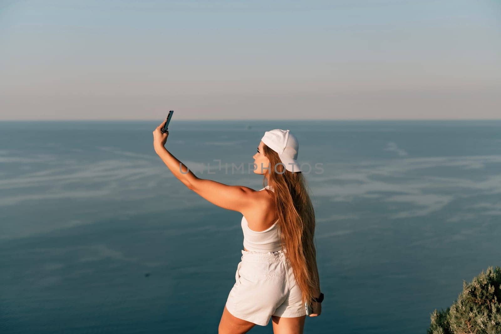 Selfie woman in a cap, white tank top and shorts makes a selfie shot mobile phone post photo social network outdoors on the background of the sea beach people vacation lifestyle travel concept. by Matiunina