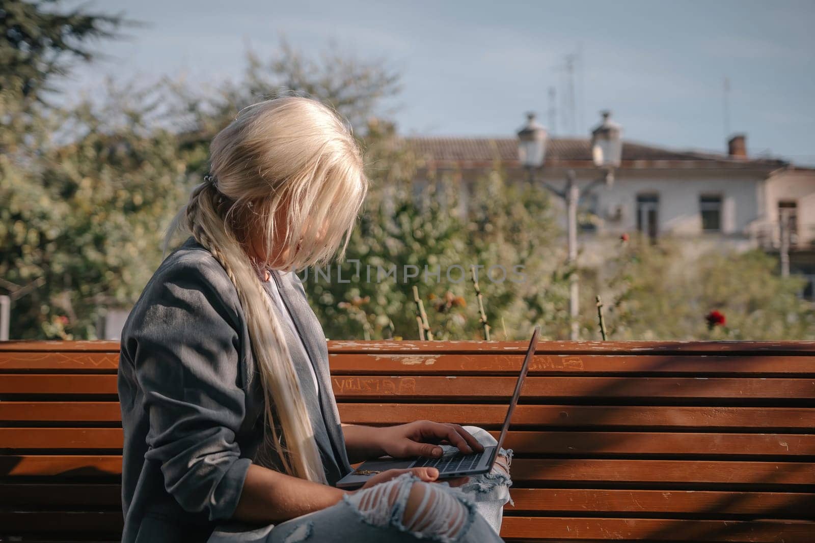 A blonde woman sits on a bench with a laptop open in front of her. She is wearing a gray jacket and has her hair in a ponytail. The scene suggests a casual and relaxed atmosphere