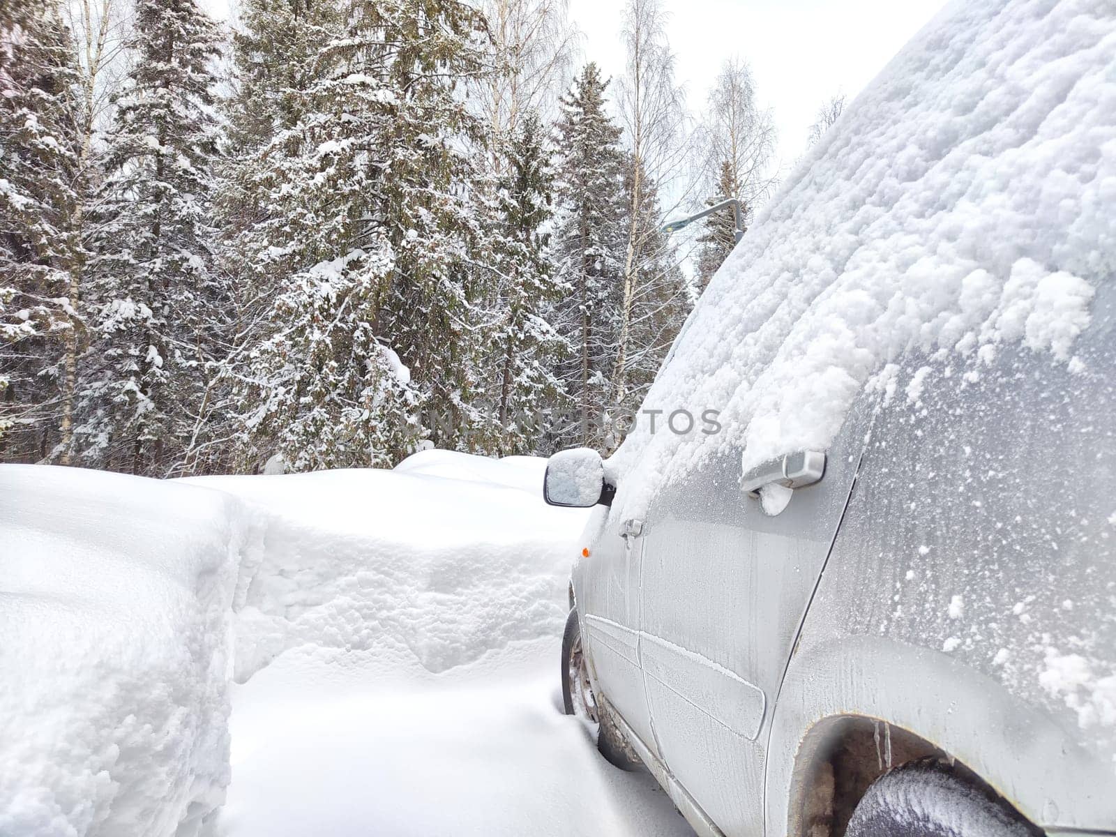 A car is almost entirely blanketed by a thick layer of fresh snow, with only a portion of the side mirror and front wheel visible. Heavy snowfall and cold weather conditions