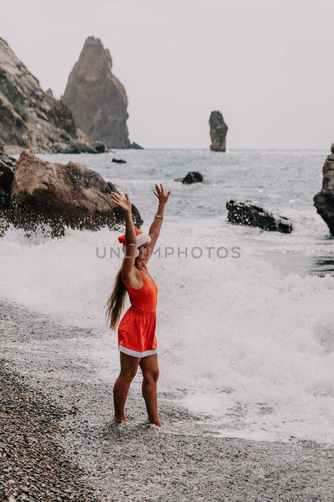 Woman summer travel sea. Happy tourist in red bikini and Santas hat enjoy taking picture outdoors for memories. Woman traveler posing on the beach surrounded by volcanic mountains, sharing travel joy by panophotograph