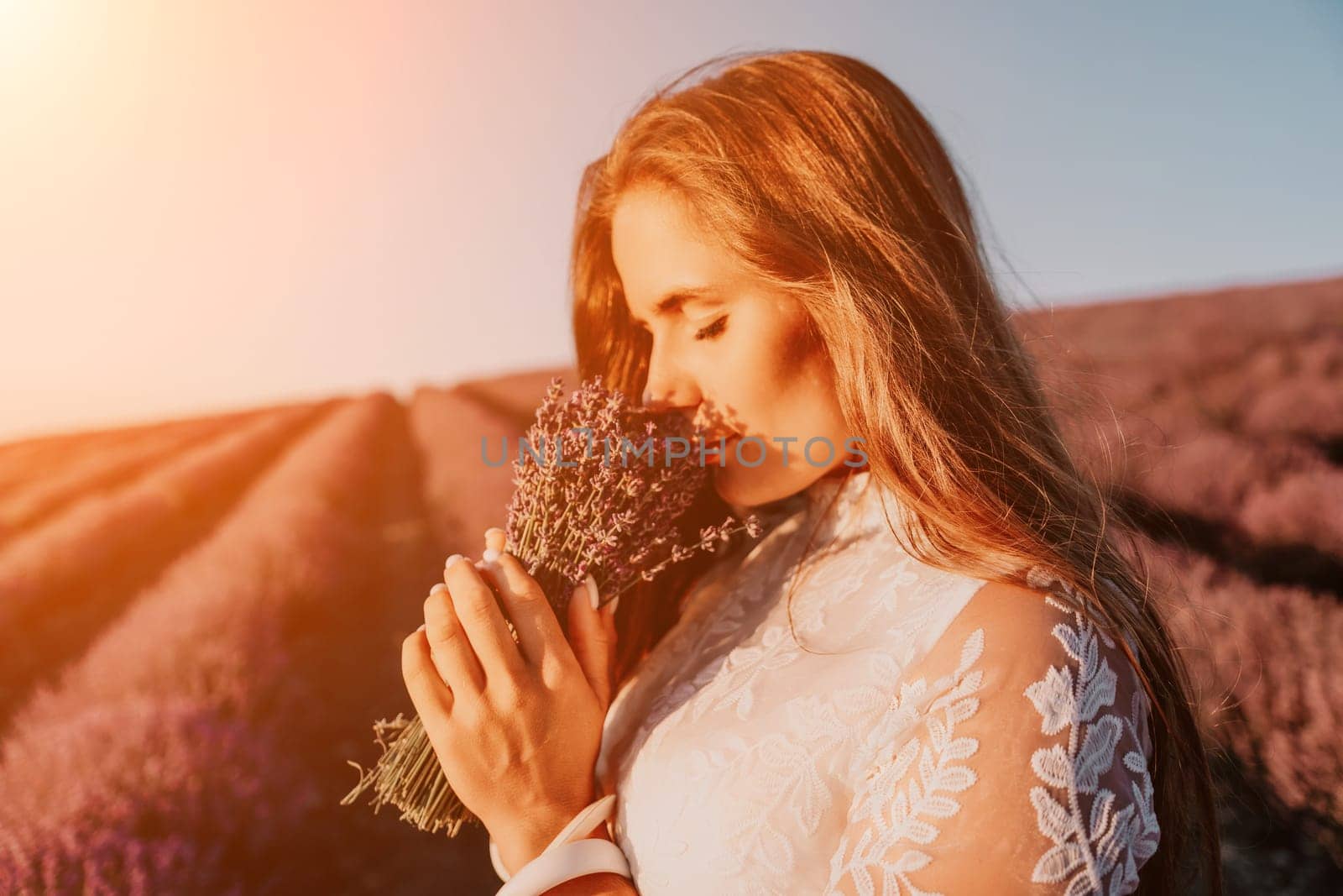 Close up portrait of young beautiful woman in a white dress and a hat is walking in the lavender field and smelling lavender bouquet.