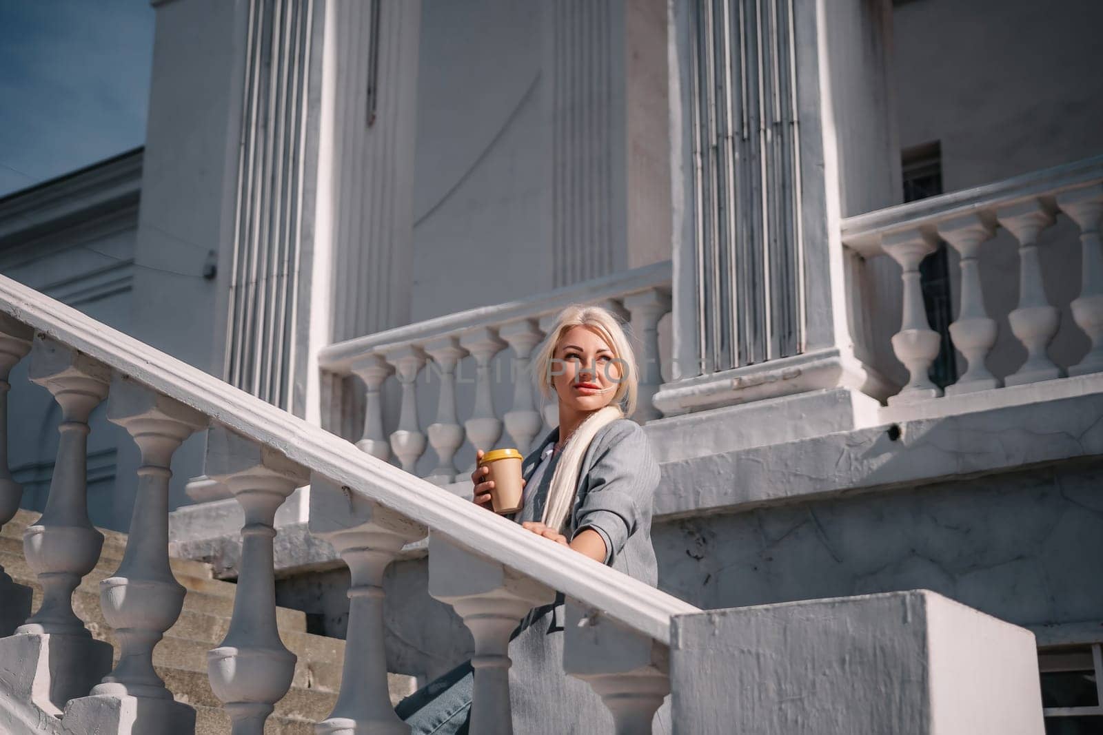 A woman is standing on a white staircase with a cup in her hand. She is smiling and looking up at the sky
