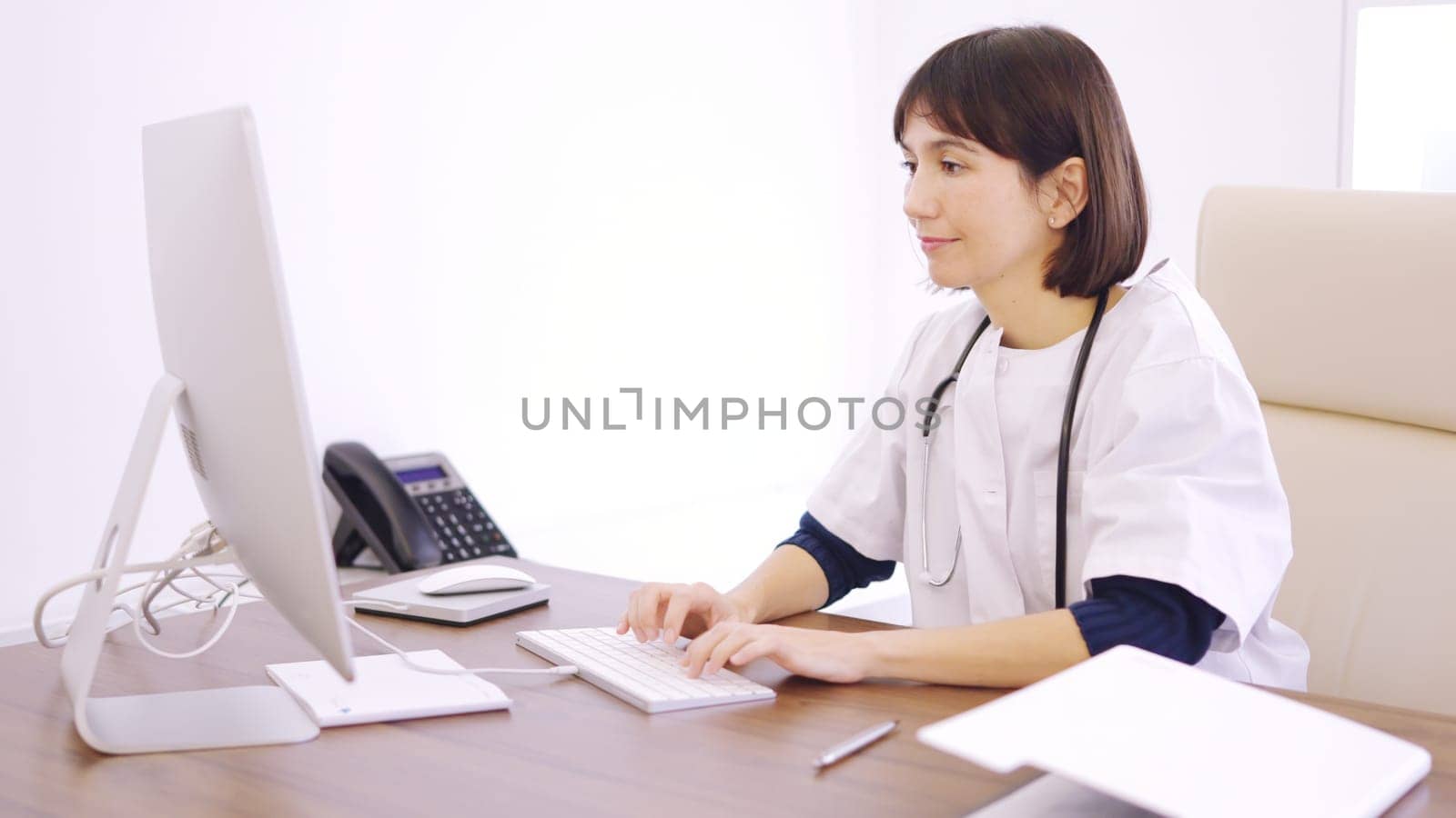 Doctor using computer sitting on a desk in a clinic by ivanmoreno