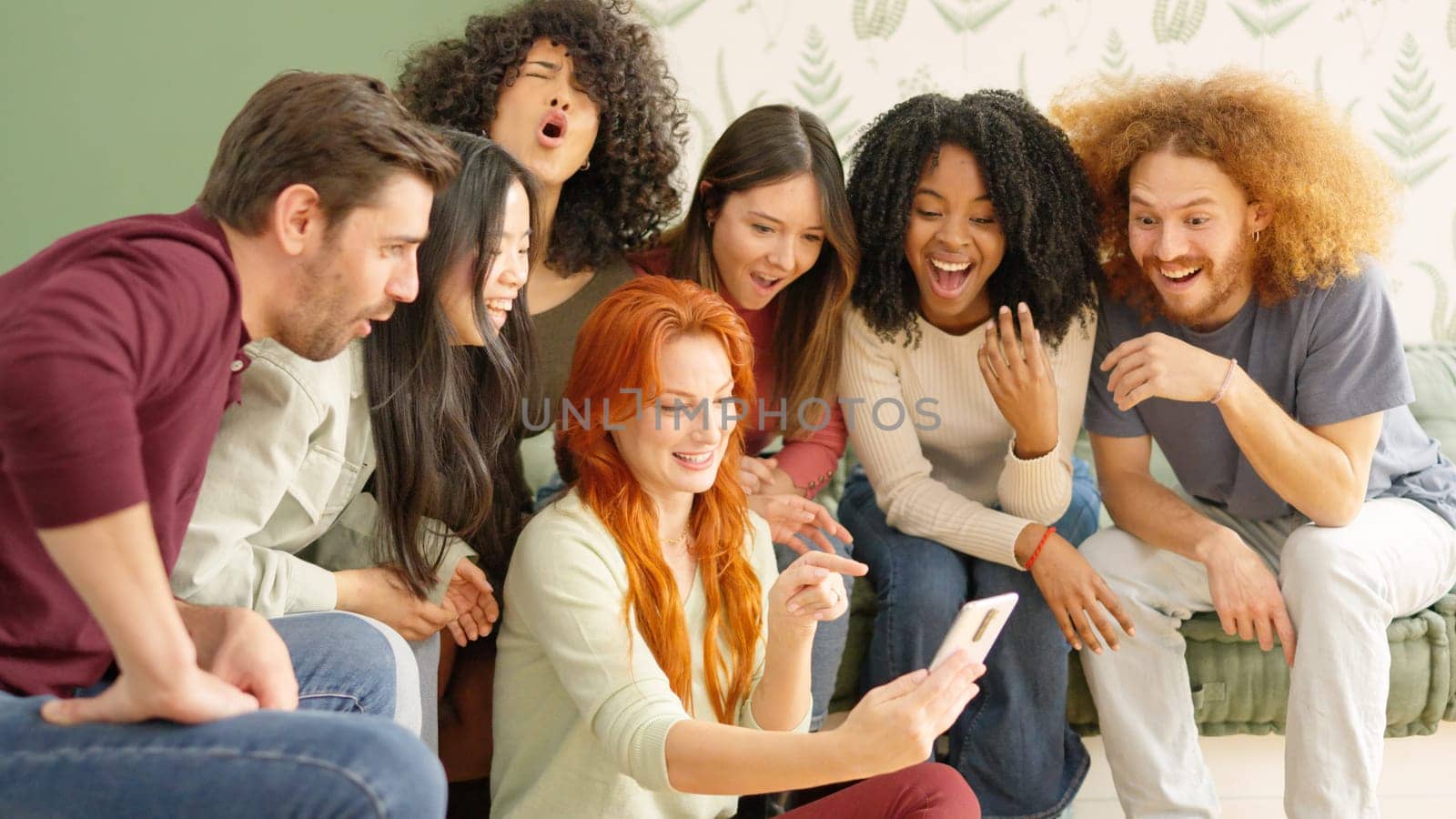 Multi-ethnic group of friends looking at a mobile of one woman and laughing at one video on it sitting on a sofa