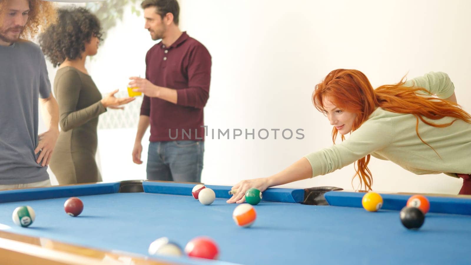 Redhead woman hitting a ball playing pool with friends at home