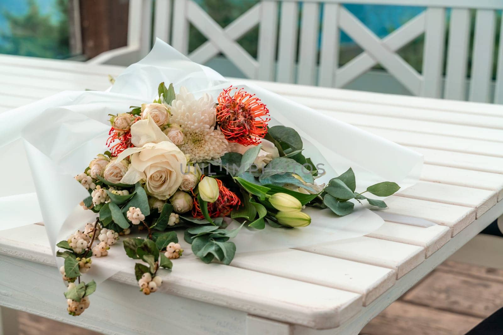 A bouquet of flowers is sitting on a table by the ocean. The flowers are white and orange, and they are arranged in a vase. The table is surrounded by chairs