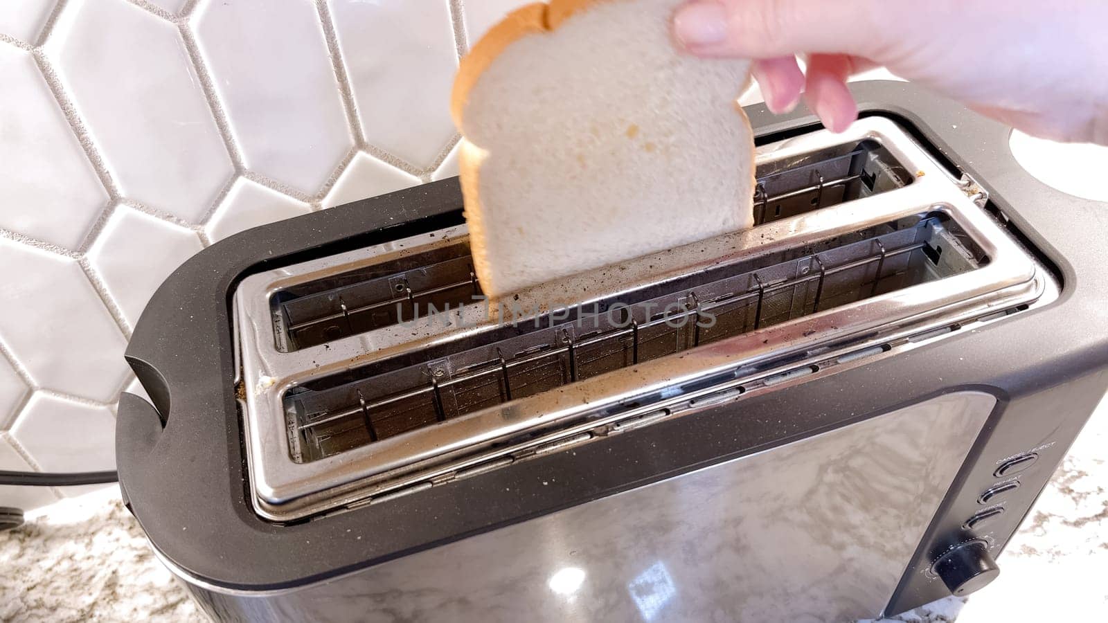 The image shows a sleek, contemporary toaster with a single slice of bread visible through its long slot, set against a tiled kitchen backsplash, with active blue indicator lights.