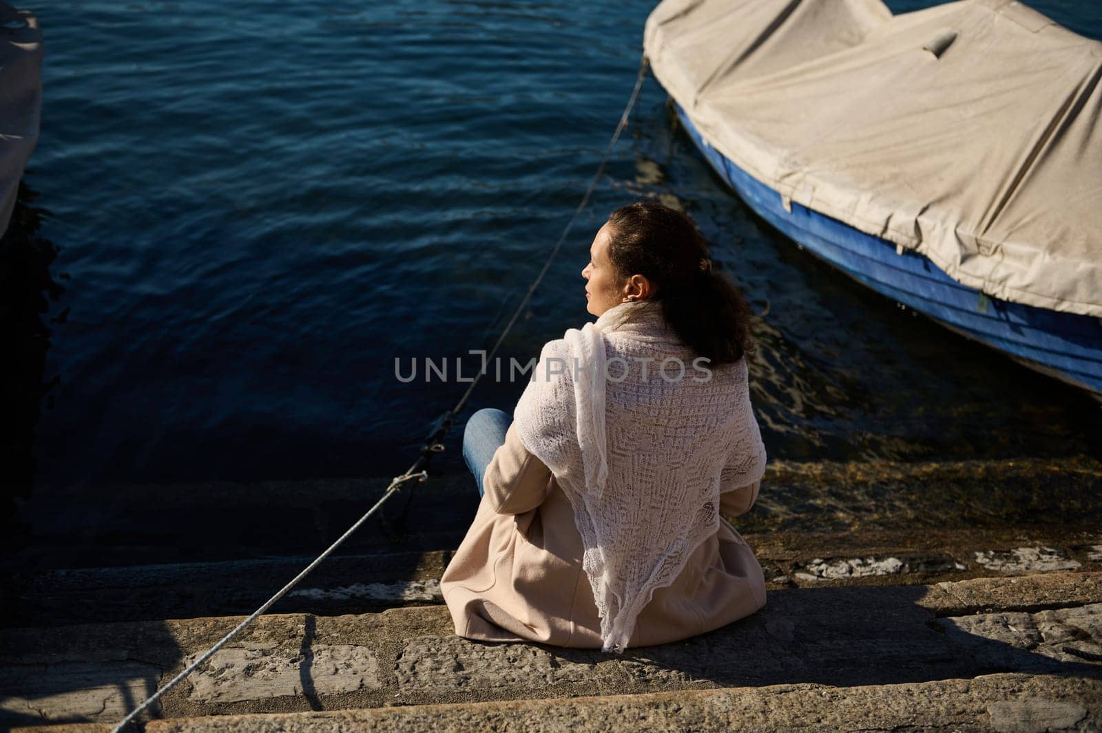 Rear view of a young woman relaxing, sitting near a small boat on the edge by lake, admiring the beautiful Alpine nature from the waterfront. Como, Milan, Lombardy, Italy. Travel and tourism concept by artgf