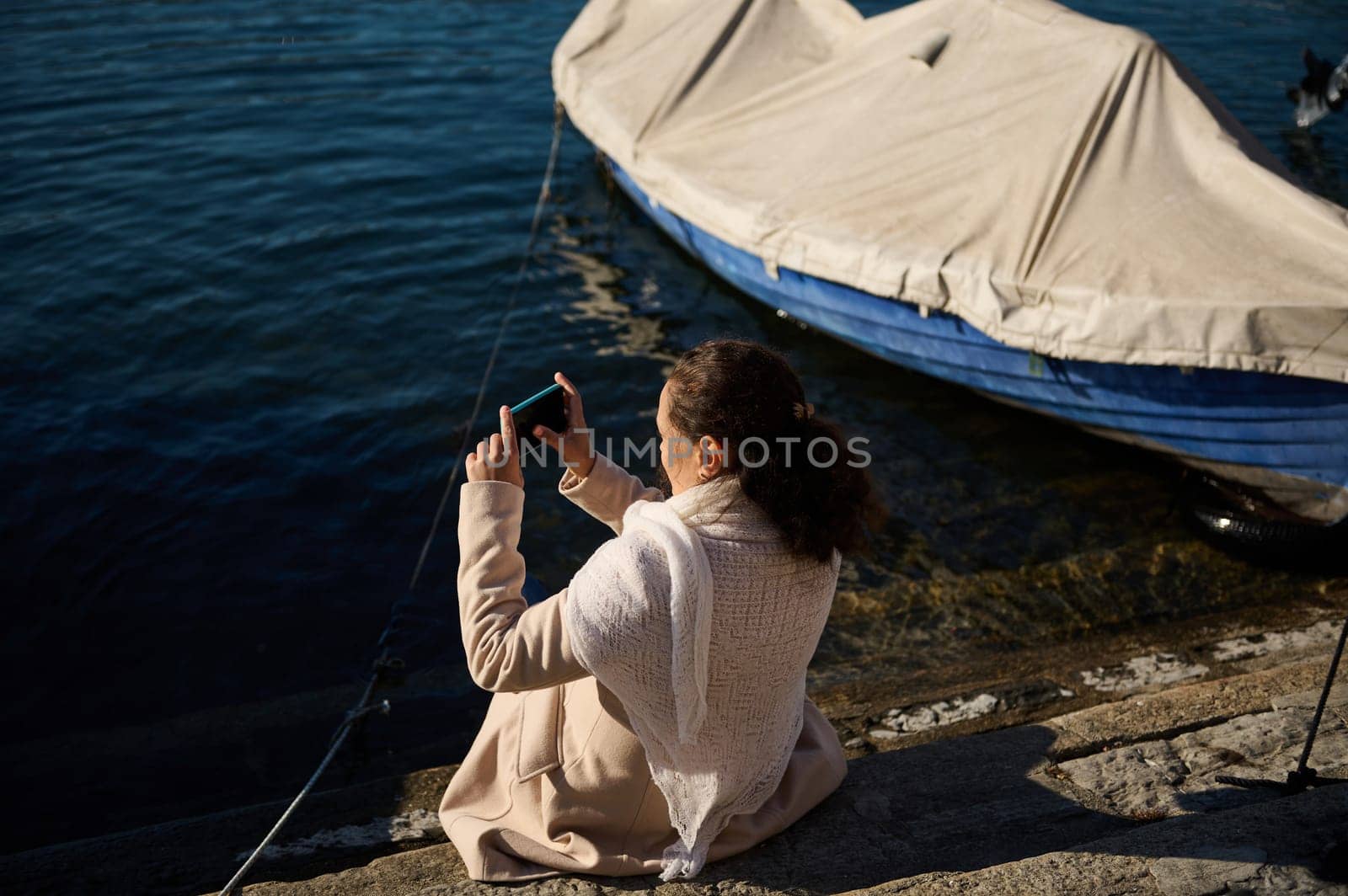 Rear view of a relaxed young woman in beige coat, sitting nears small boats by the Como lake and photographing the landscape on her smart mobile phone. by artgf