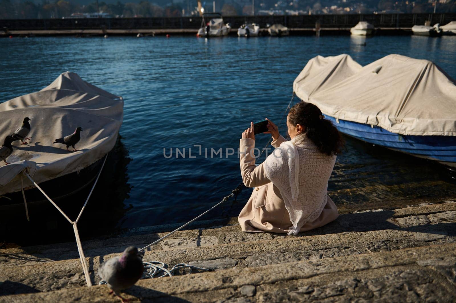 Rear view of a young woman in beige coat, sitting nears boats by the Como lake and photographing the landscape on her smart mobile phone. Pretty female tourist relaxing in the lakefront by artgf