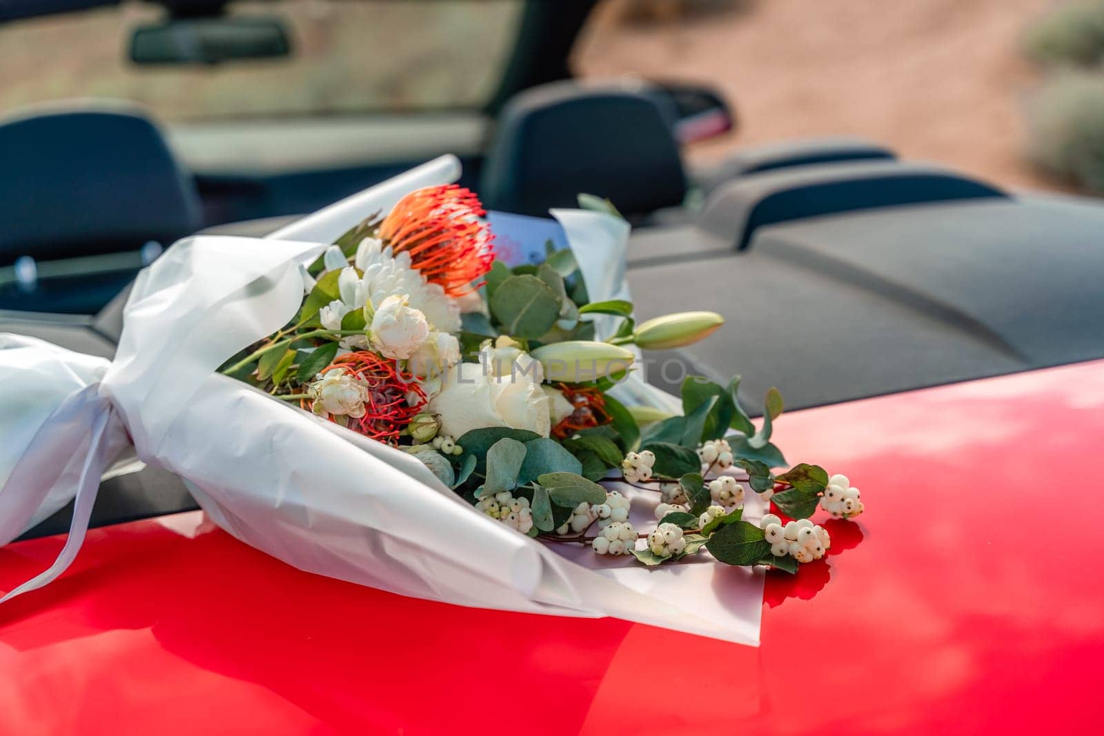 A bouquet of flowers is sitting on the hood of a red car. The flowers are white and red, and they are arranged in a vase. Concept of romance and love