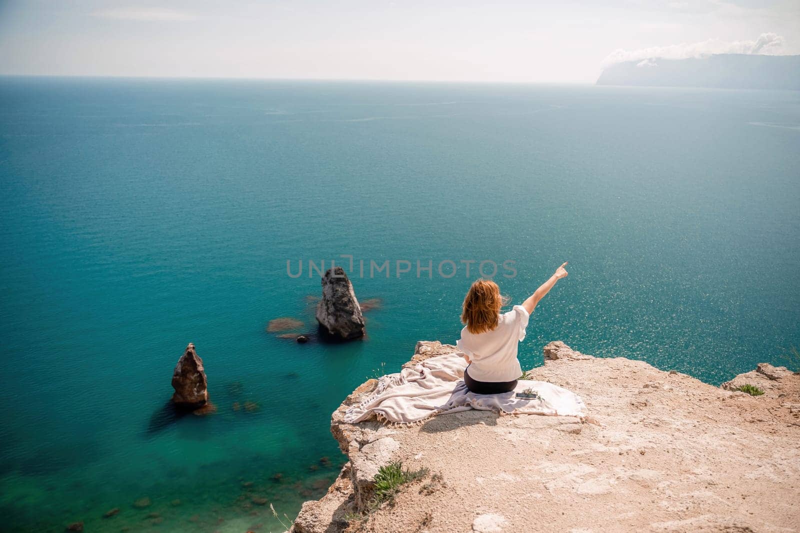 A woman is sitting on a rock overlooking the ocean. She is pointing to the water. The scene is peaceful and serene, with the woman enjoying the view and the calming sound of the waves