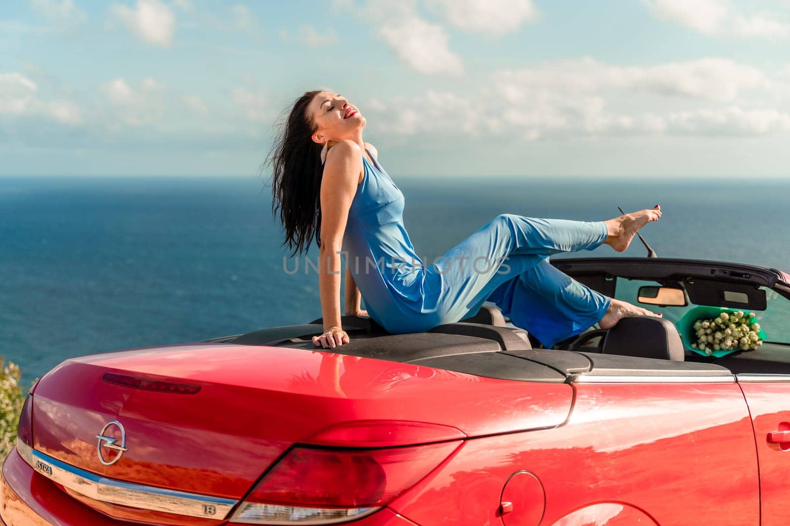 A woman is sitting in a red convertible car, smiling and enjoying the view of the ocean. Scene is happy and relaxed