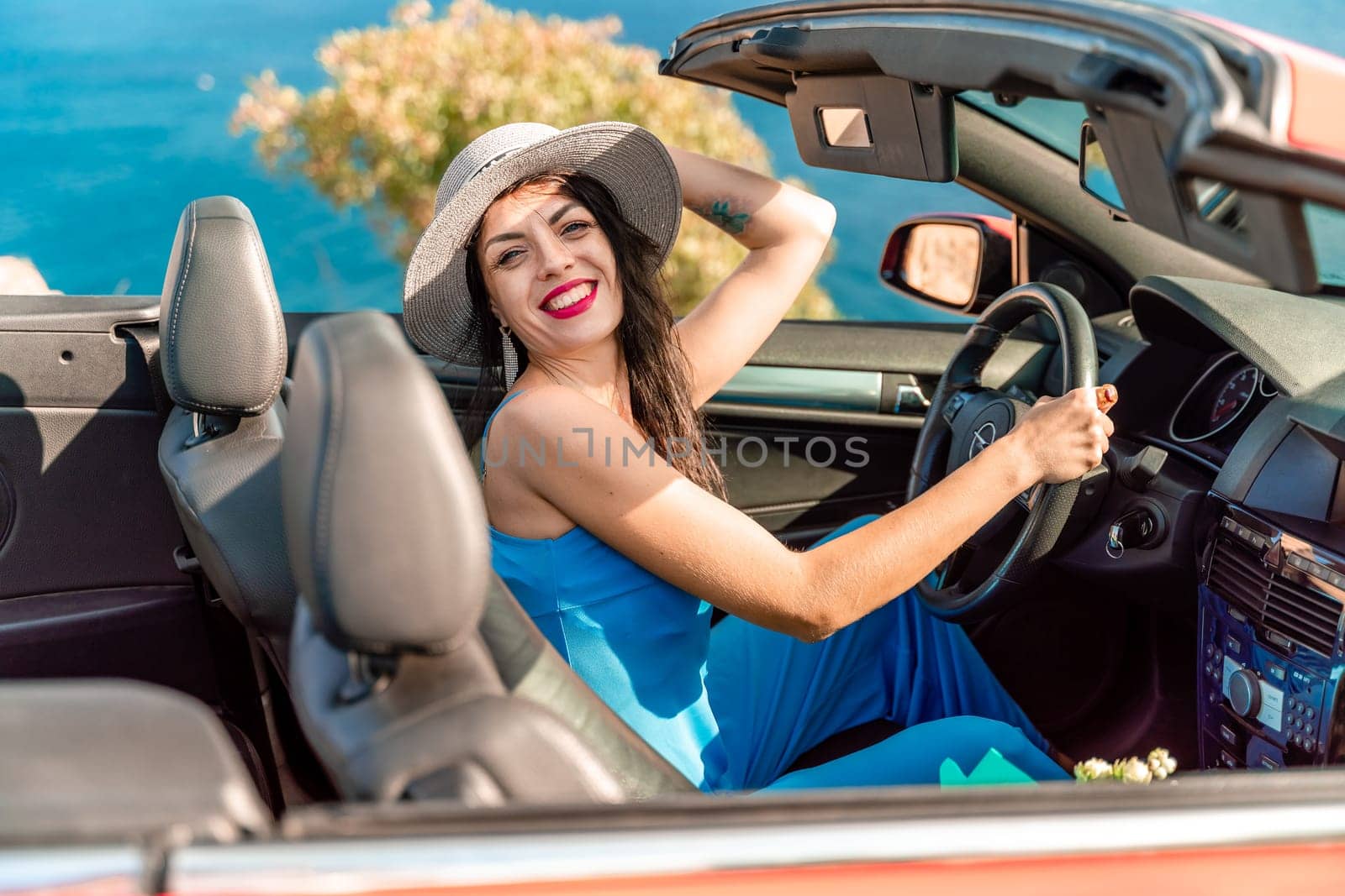 A woman is driving a red convertible with a hat on. She is smiling and she is enjoying the ride