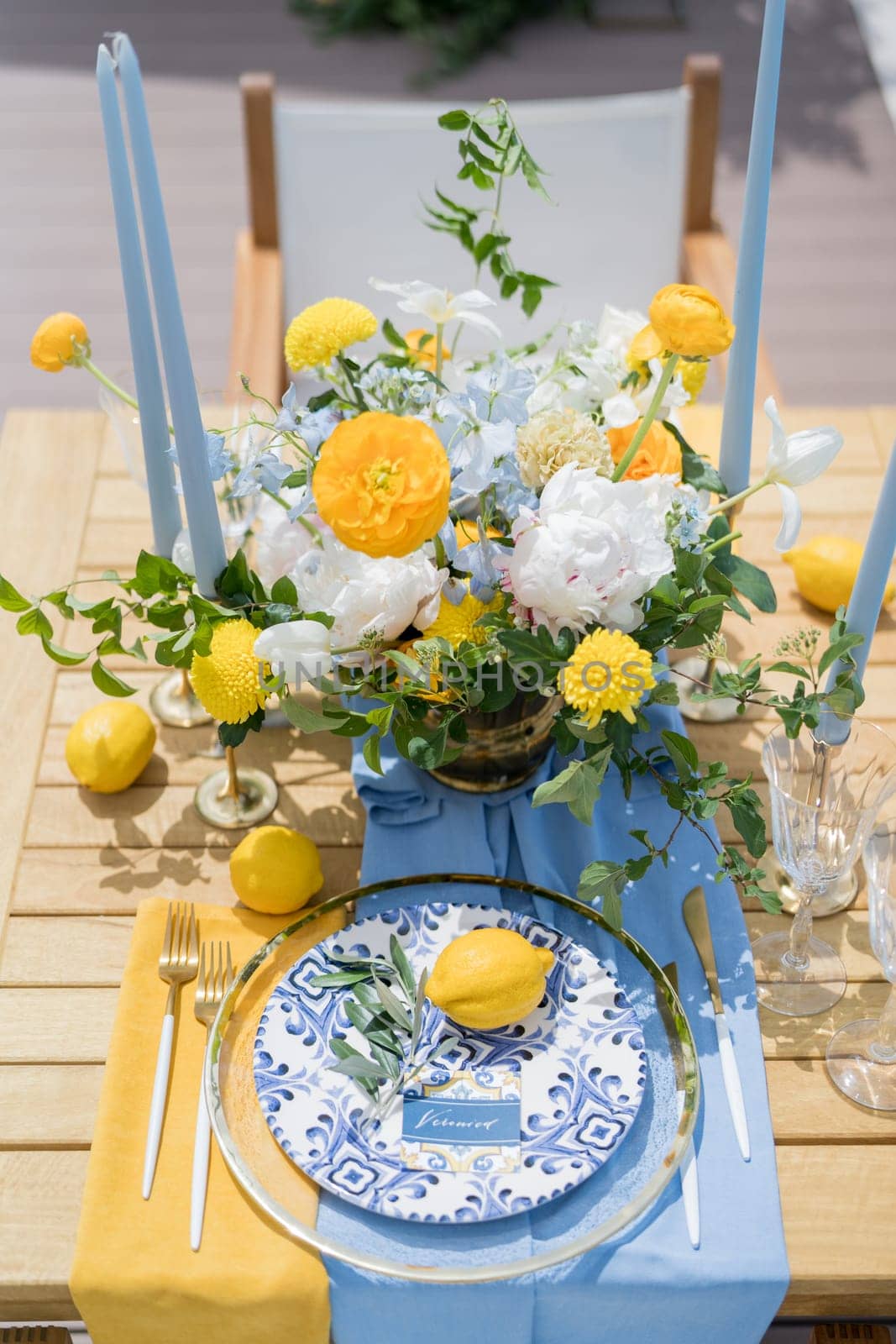 Name card lies next to an olive branch and a lemon on a plate on a set table near a colorful bouquet of flowers. High quality photo