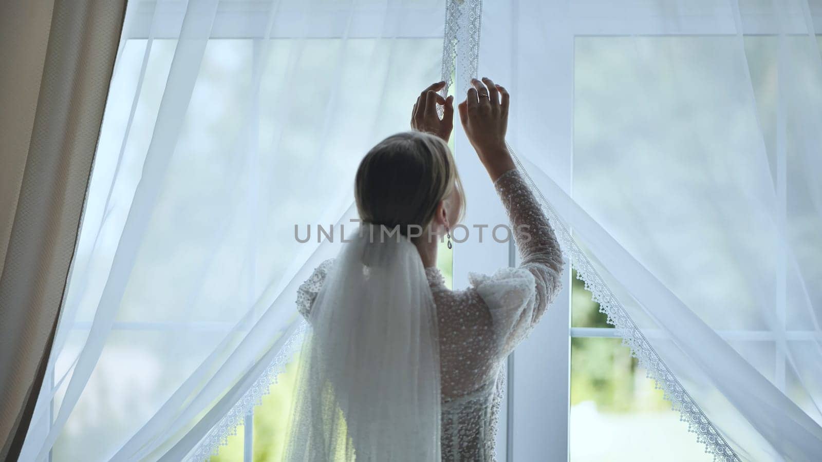 The bride adjusts the curtains at the window. by DovidPro