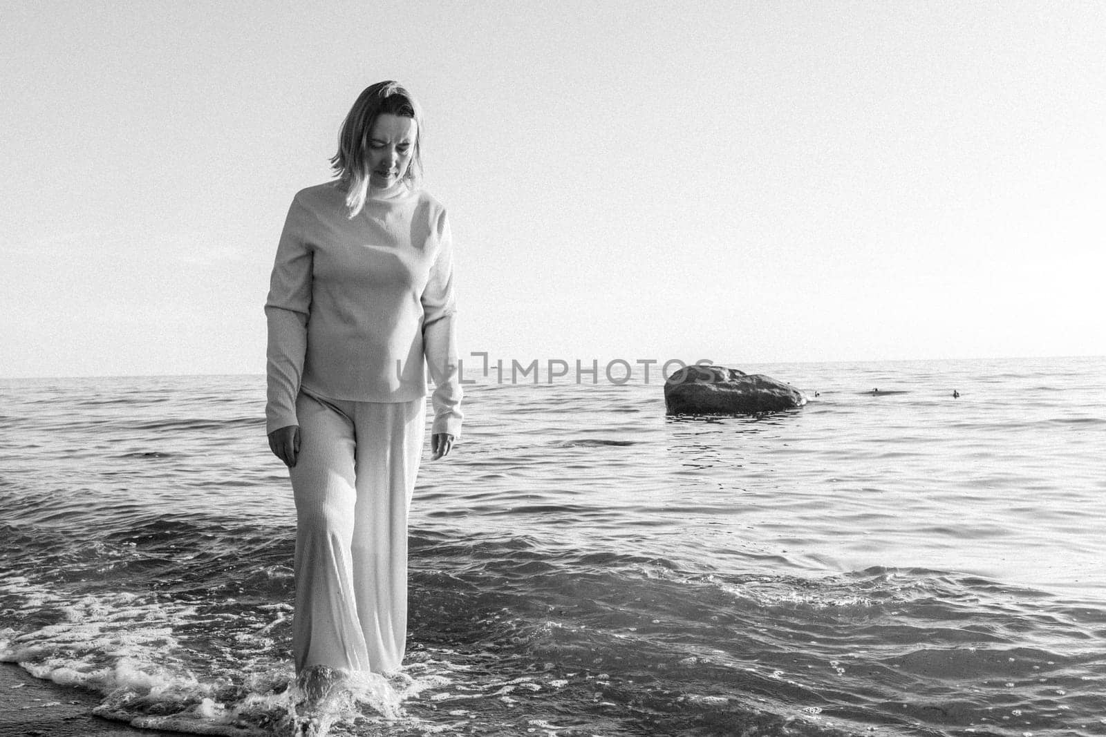 A woman standing waist-deep in the water at a sandy beach, with waves gently splashing around her. Black and white photography.