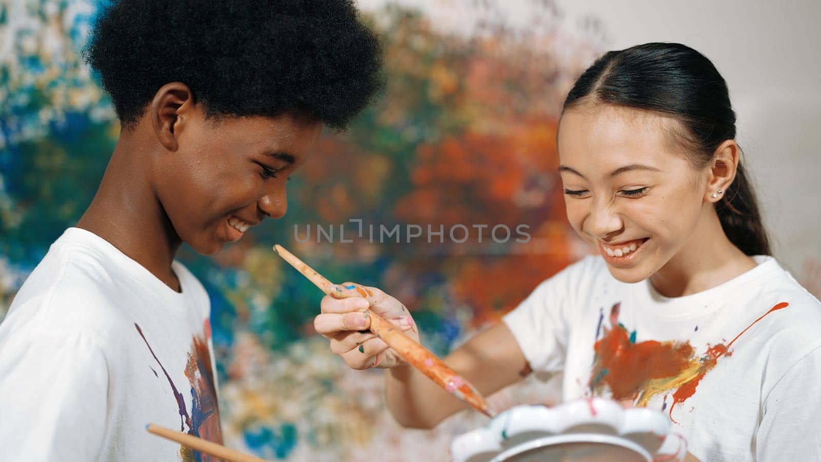 Smiling diverse children paint color on each other shirt at wall. Edification. by biancoblue