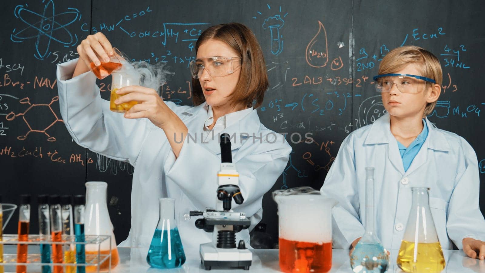 Smart caucasian teacher mixing chemical liquid in science laboratory. Professional instructor doing experiment in STEM science class with microscope at blackboard written chemical theory. Erudition.