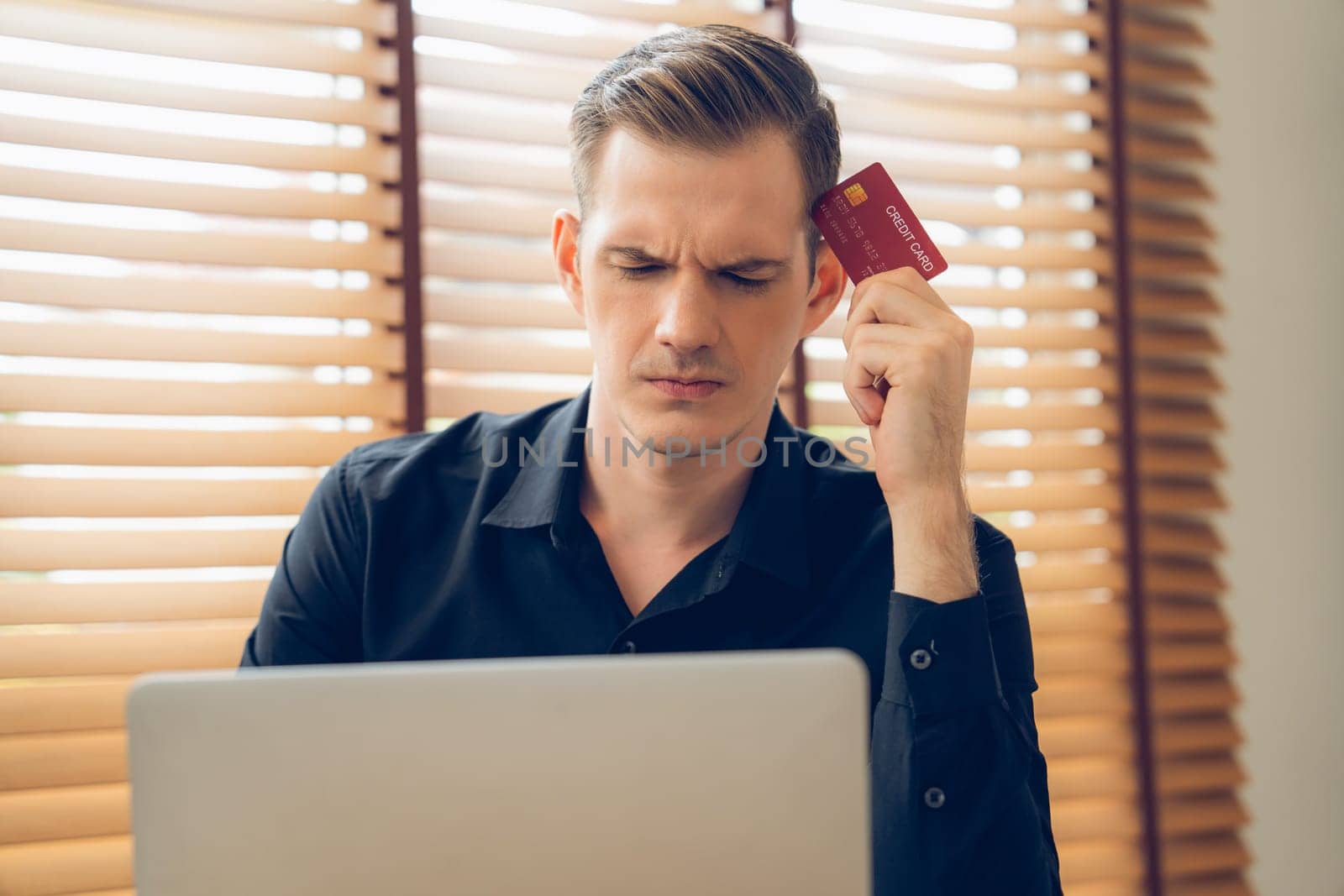 Stress young man sit at table, as he pay bills online with credit card from financial debt problem, monthly expense and credit card debt. Over spending money lifestyle problem. Unveiling