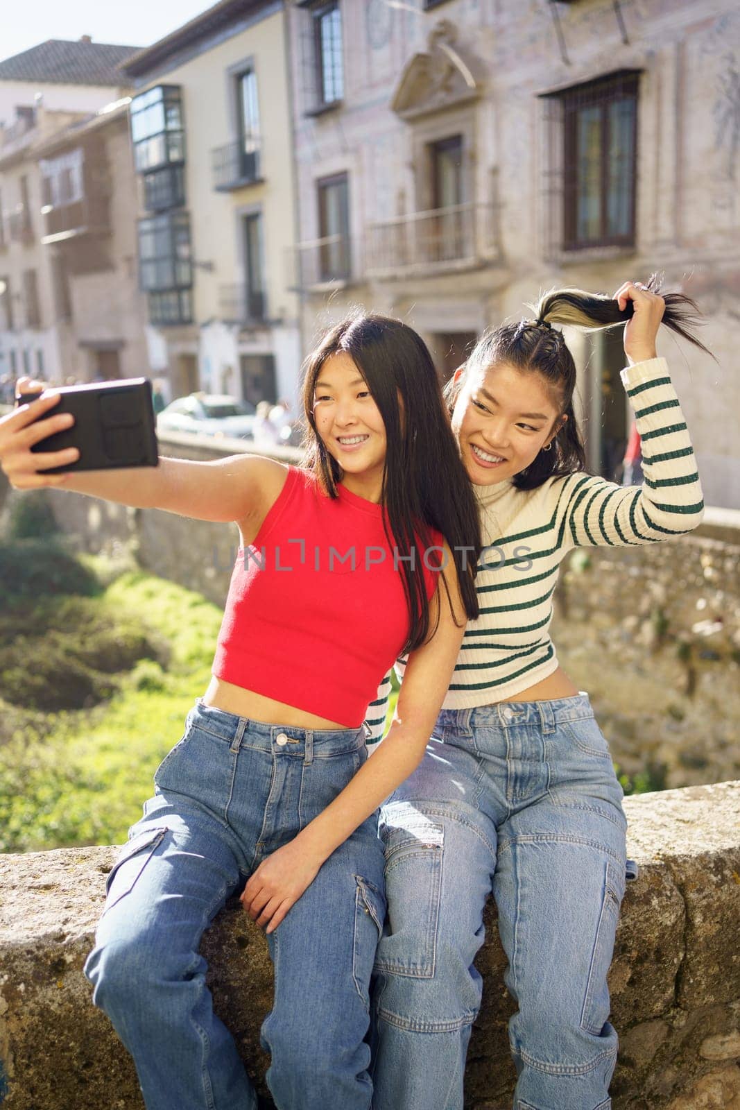 Cheerful Asian female friends sitting on stone and taking self portrait while enjoying time together in Granada