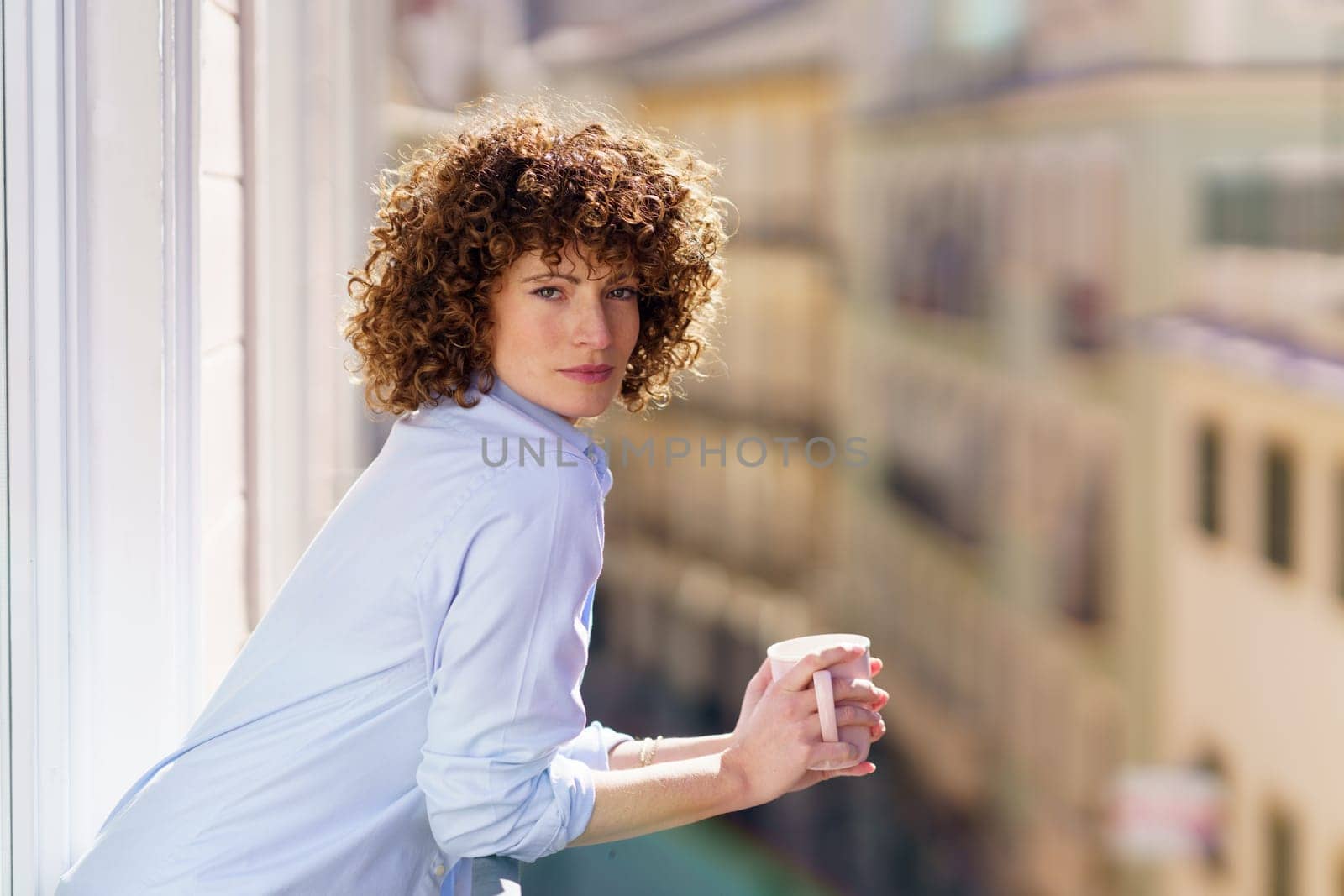 Adult woman with brown curly hair leaning on railing of balcony while enjoying cup of hot beverage against blurred background