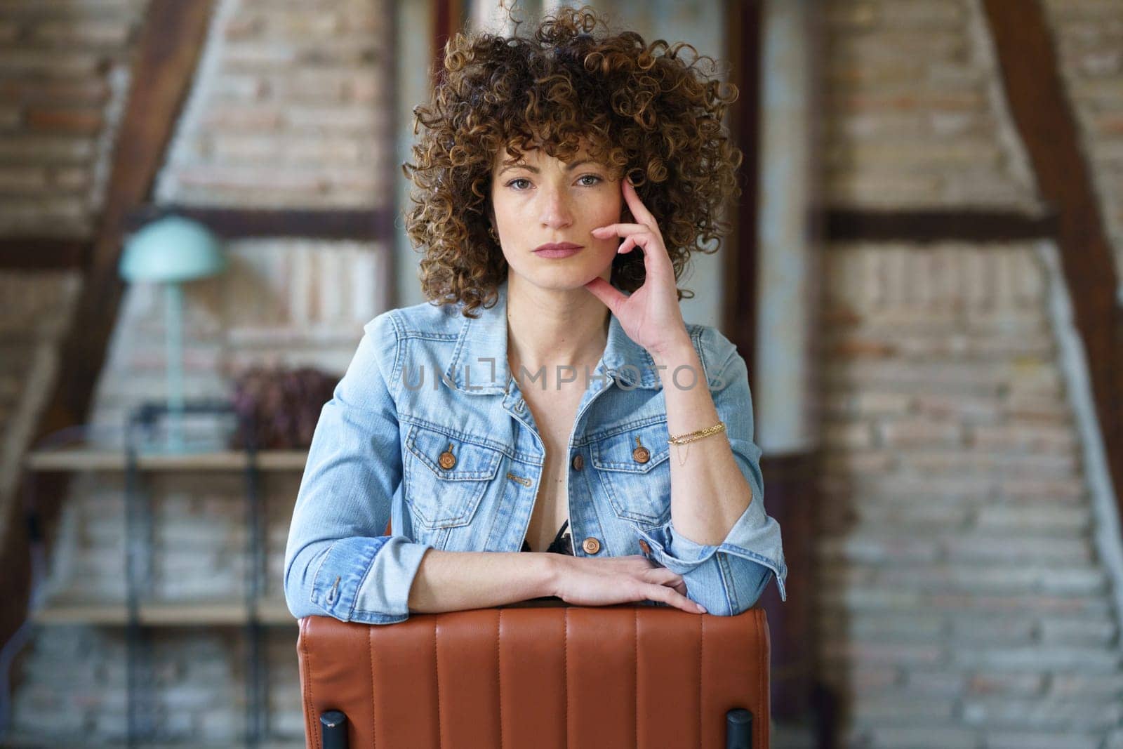 Serious young woman wearing denim jacket sitting on leather chair and touching hair while looking at camera against blurred brick wall