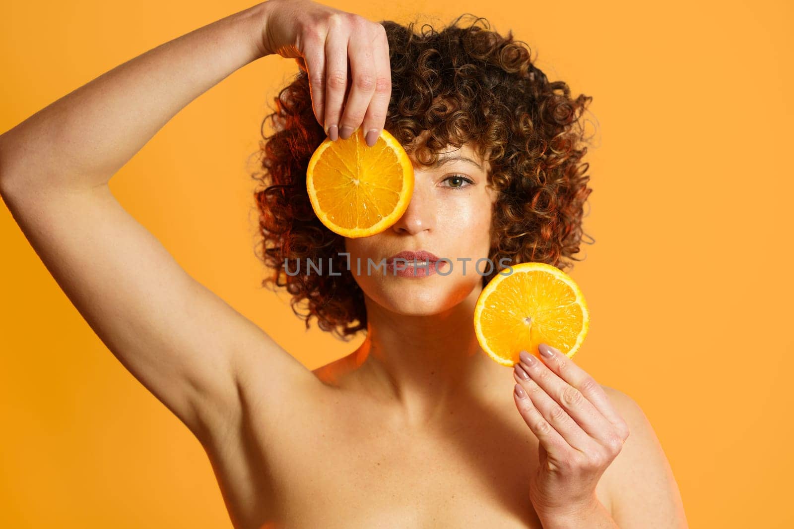 Serious woman with curly hair covering face with slices of ripe citrus during beauty routine against orange background