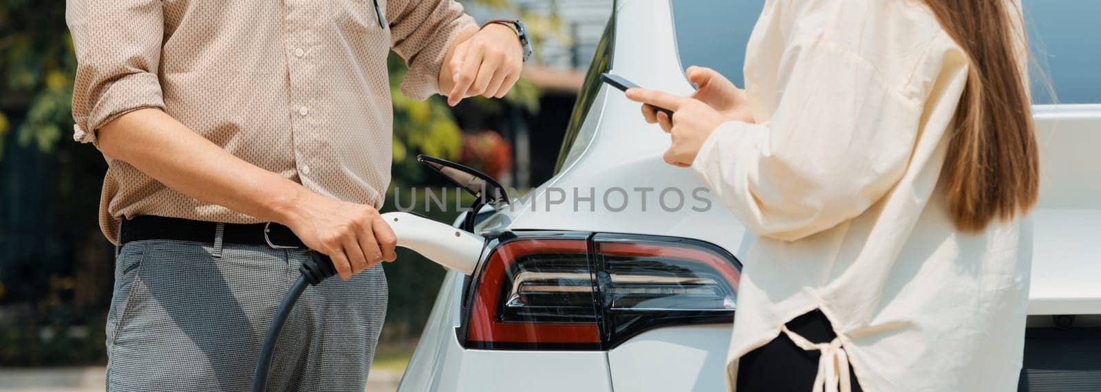 Young couple use smartphone to pay for electricity at public EV car charging station green city park. Modern environmental and sustainable urban lifestyle with EV vehicle. Panorama Expedient