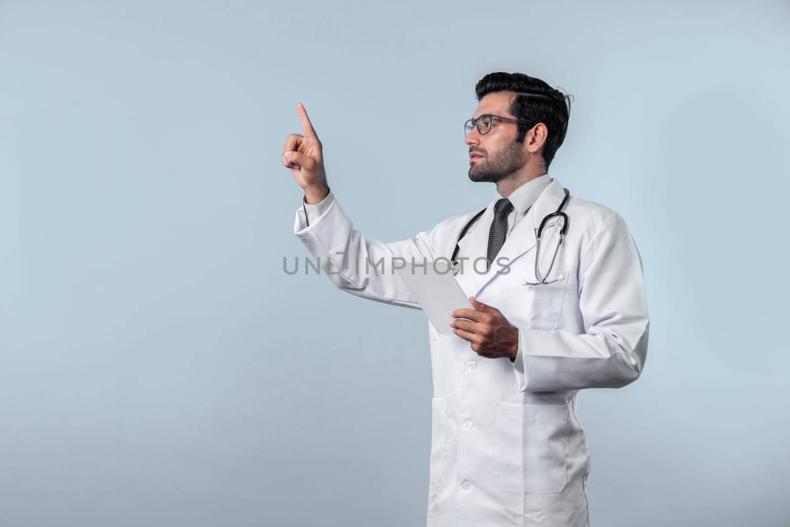 Caucasian smart doctor pointing finger while standing at white background and wearing lab coat. Skilled specialist doctor using medical technology while pointing and holding health data. Deviation.