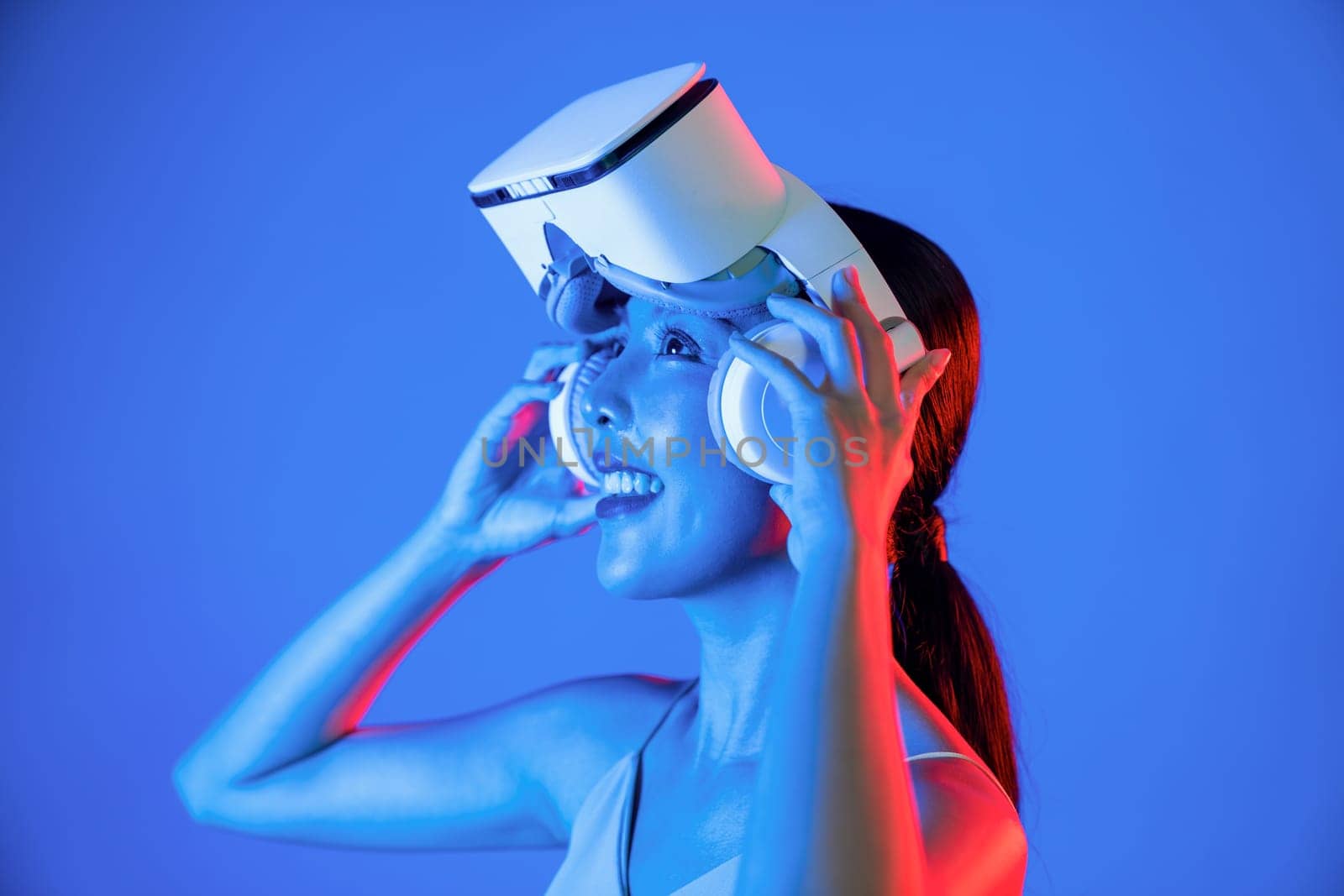 Smart female stand hit by neon light strapping VR headset above eye.The gadget function for connecting metaverse, future cyberspace technology. Woman raise arms holding goggles by hand. Hallucination.