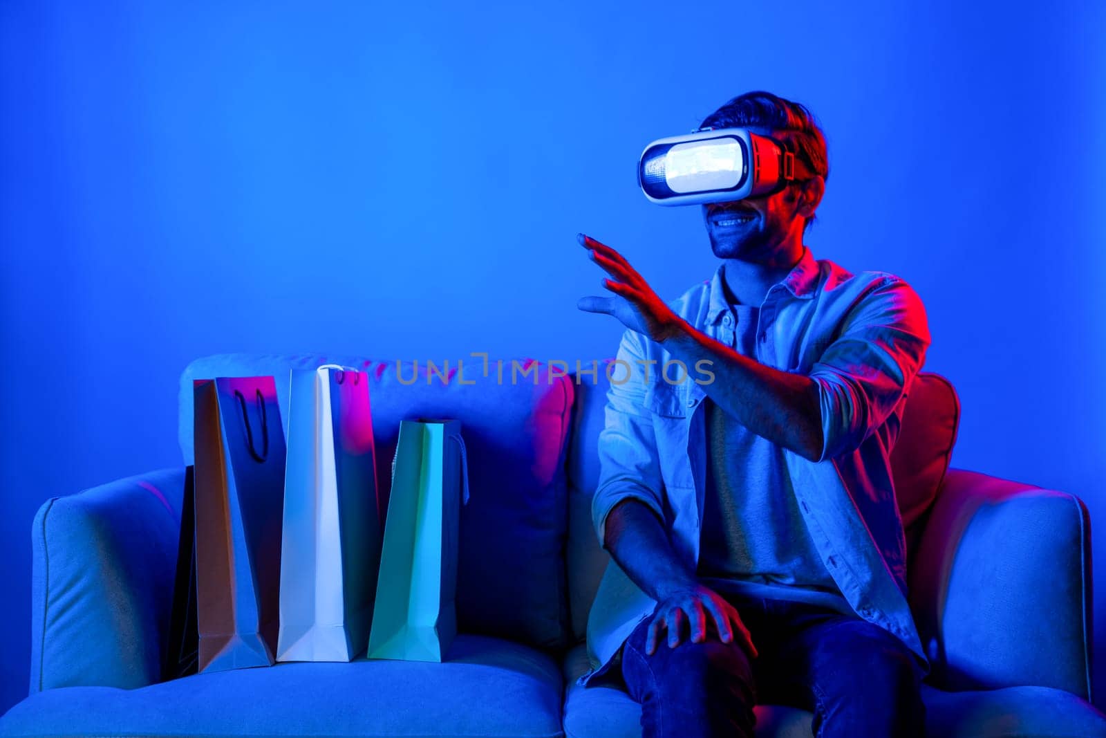 Caucasian man with vr glass posing pointing finger while sitting at sofa with shopping bags. Person selecting and doing shopping online by using VR technology and virtual goggle at home. Deviation.