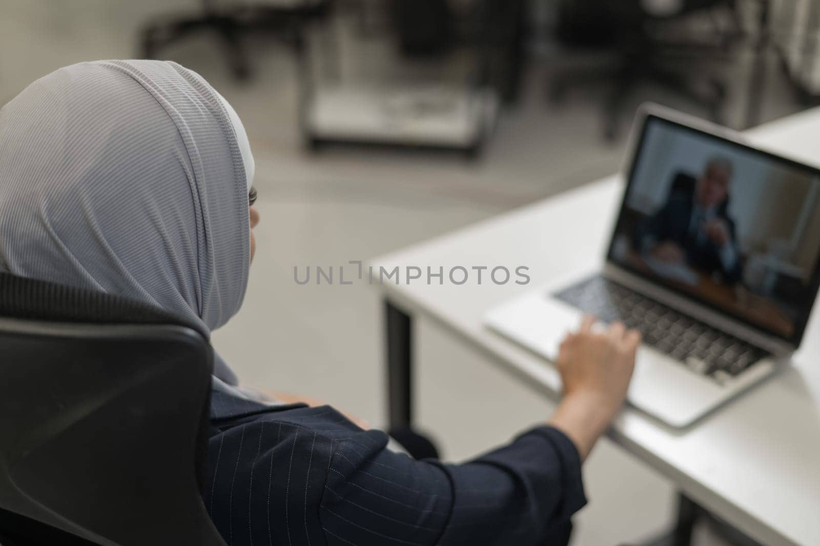 A pregnant woman in a hijab communicates with a colleague via video conference on a laptop. by mrwed54