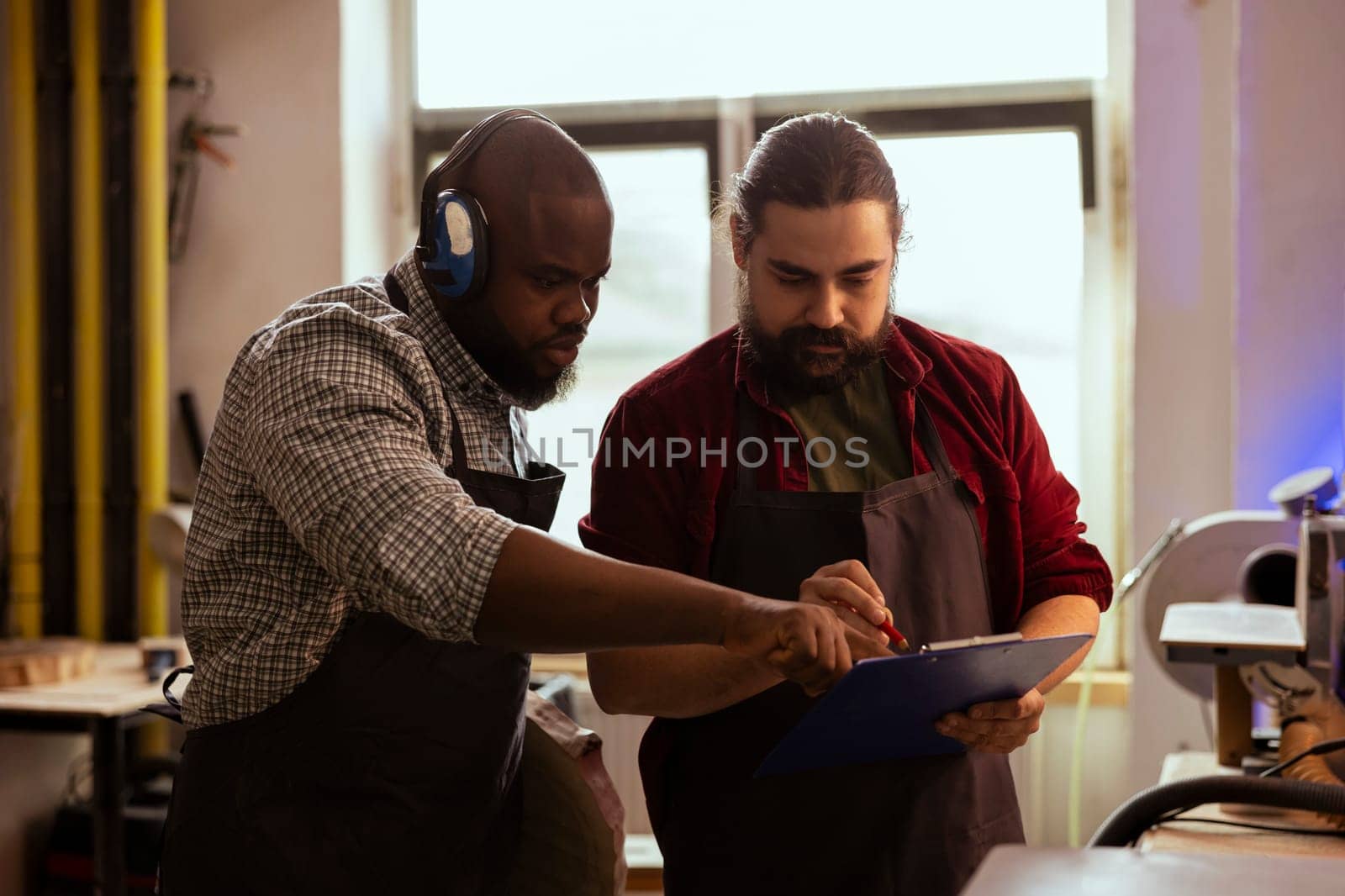 Carpenter and coworker looking over technical drawings on notepad to make creative wood art pieces. Artisan and african american man looking at blueprints to execute woodworking projects