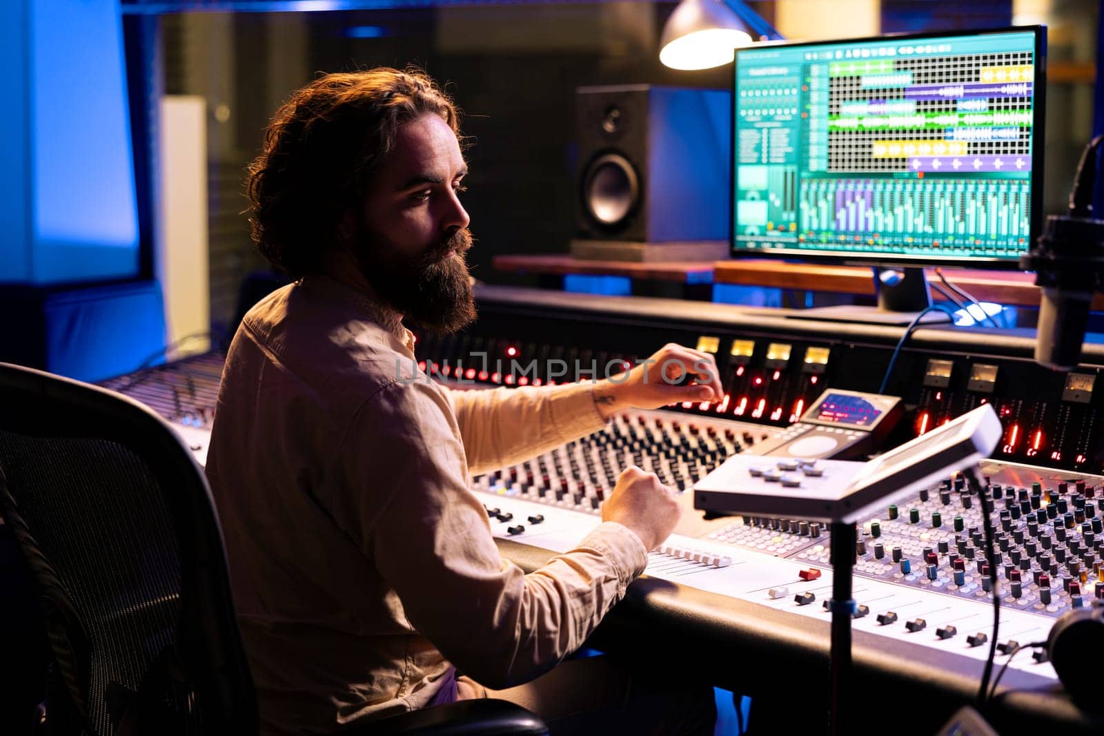 Music technician mixing and mastering songs with motorized faders and knobs, using control desk dashboard and computer. Young sound engineer creating tracks for an album in studio.