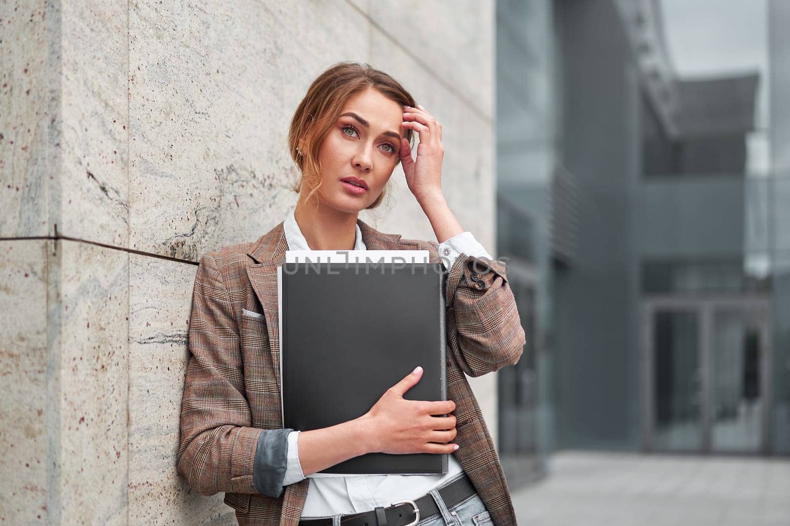 stressed businesswoman touching head standing outside business district. Worried female business person with headache pain holding document folder in hand