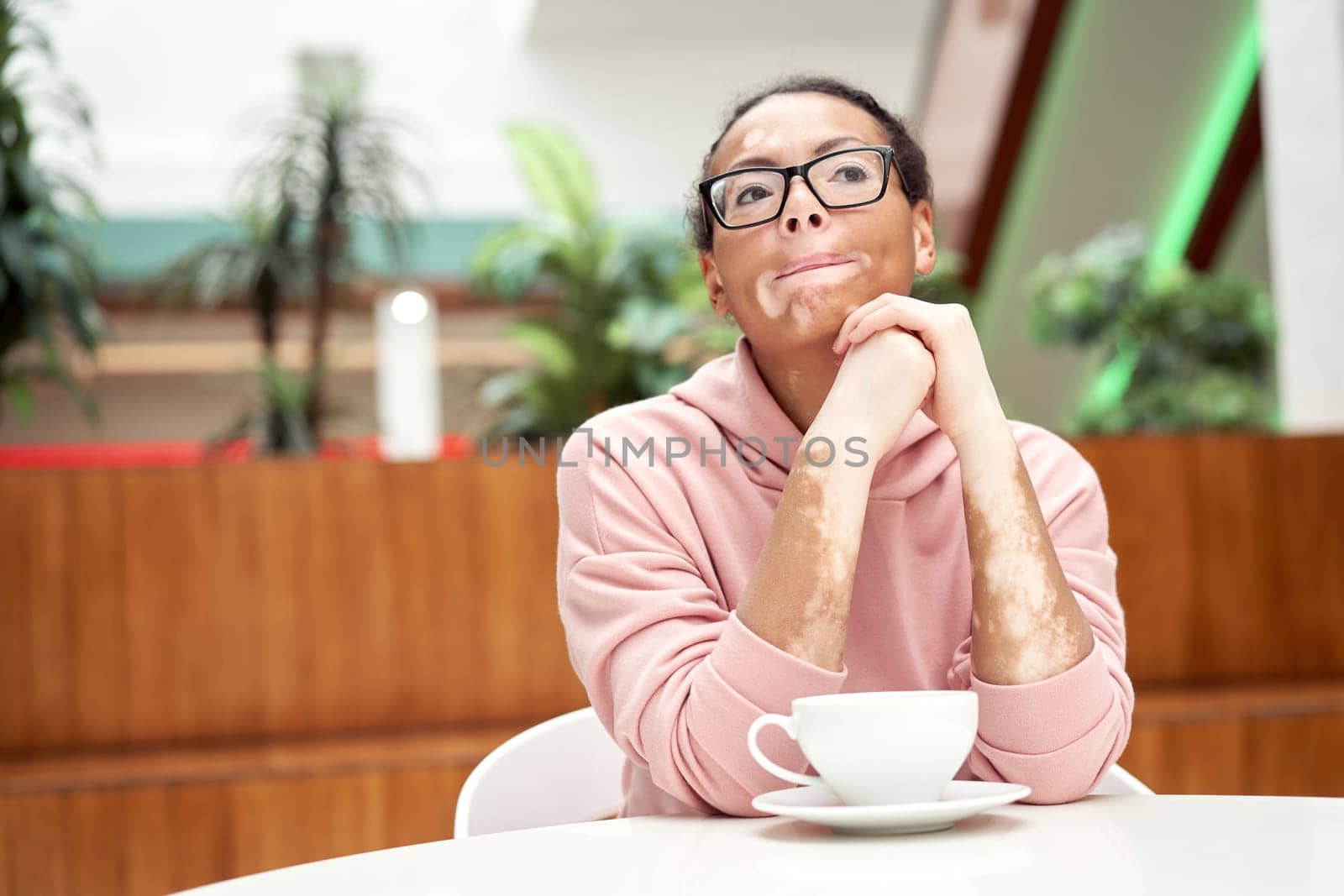 Black african american woman with vitiligo pigmentation skin problem indoor dressed pink hoodie glasses sitiing table indoor drink tea thought about something