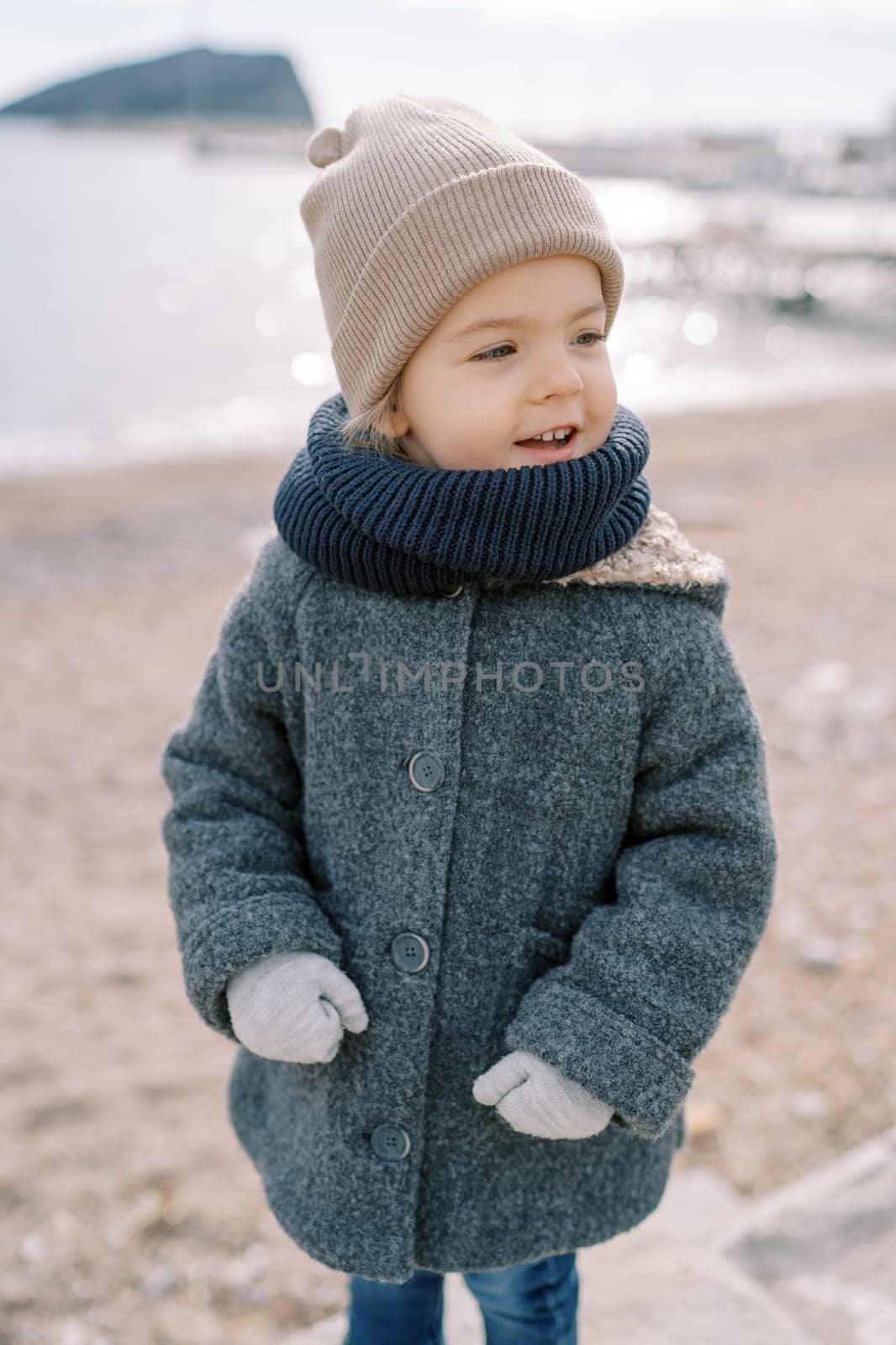 Little smiling girl in mittens stands on the beach looking to the side. High quality photo