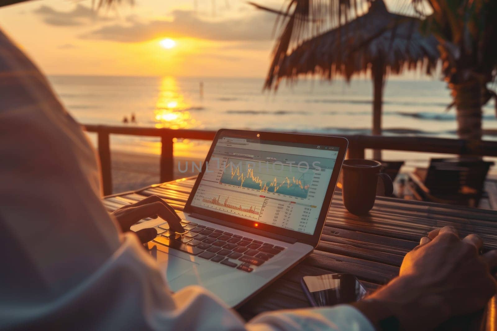 A man is sitting at a table on a beach, working on his laptop. The laptop is open to a screen displaying financial data. The man is wearing a white shirt and he is focused on his work