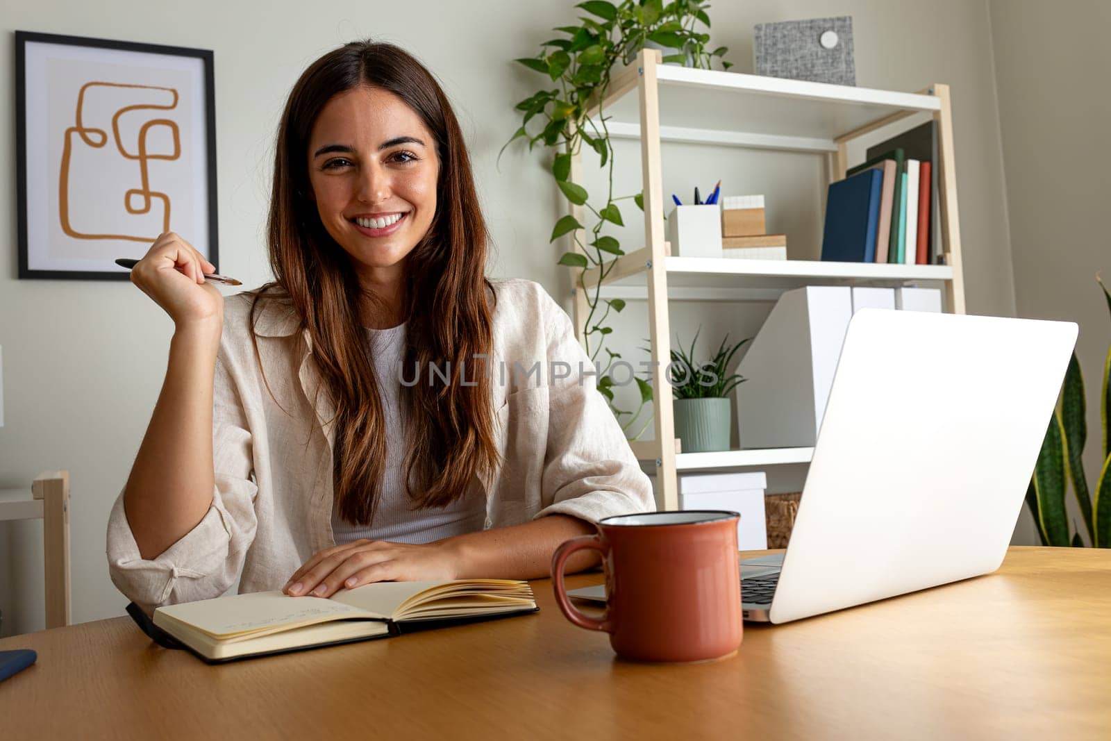 Young happy female caucasian entrepreneur handwriting on a notebook working at home office looking at camera. by Hoverstock