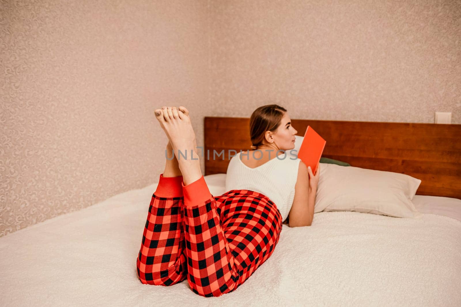 Balancing work, studies, and reading at home: Woman in red checkered pants lying on her bed with a red book and notes . by panophotograph