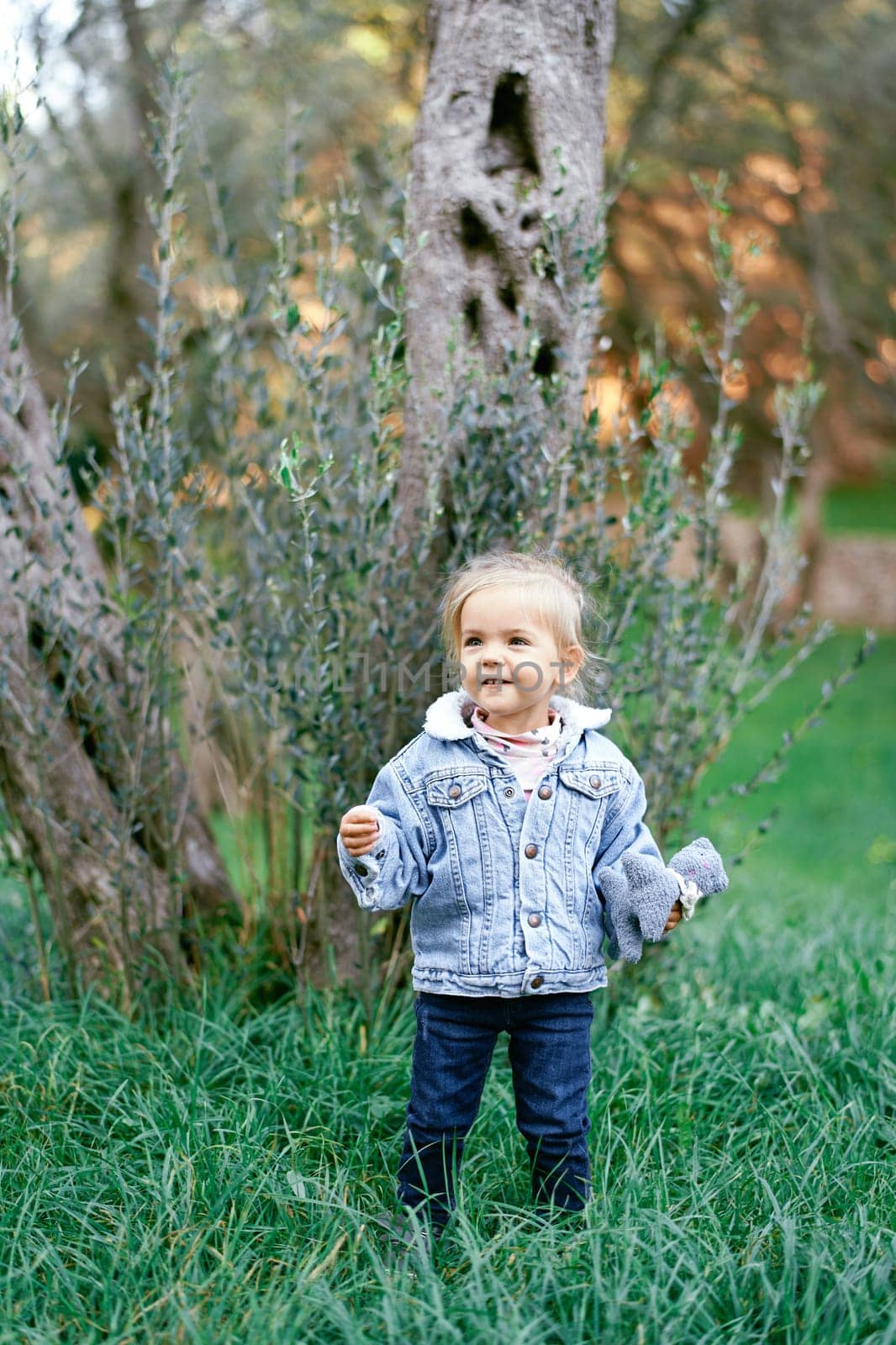 Smiling little girl stands in the grass near the tree with a soft toy in her hand. High quality photo